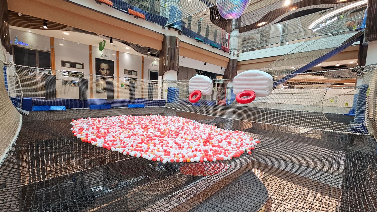 A large indoor play area with a netted floor filled with red and white plastic balls in the centre. Two inflatable white structures with red rings are suspended above the net. The background features a multi-level shopping centre with glass railings, columns, and various shopfronts. The ceiling has modern lighting fixtures, and the floor beneath the net is visible through the mesh.