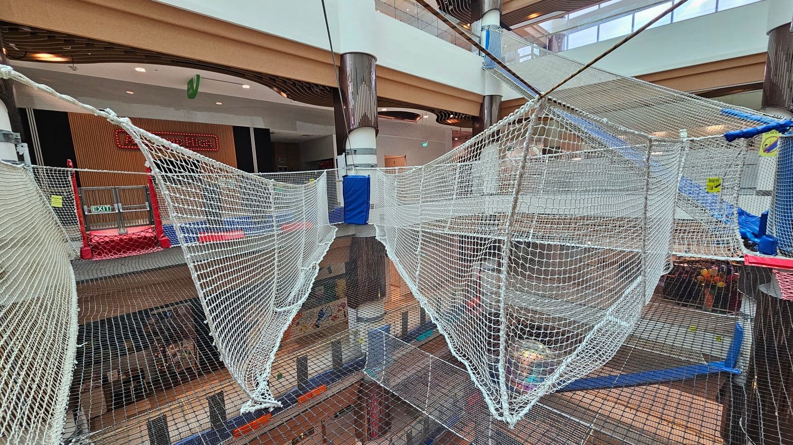 A large indoor play area with suspended net walkways and platforms. The nets are white and form a pathway across multiple levels. In the background, there are wooden panels and a red exit sign. The area is supported by large cylindrical columns. The floor below is visible through the nets, showing a lower level with various activities. The ceiling has a modern design with recessed lighting and skylights.