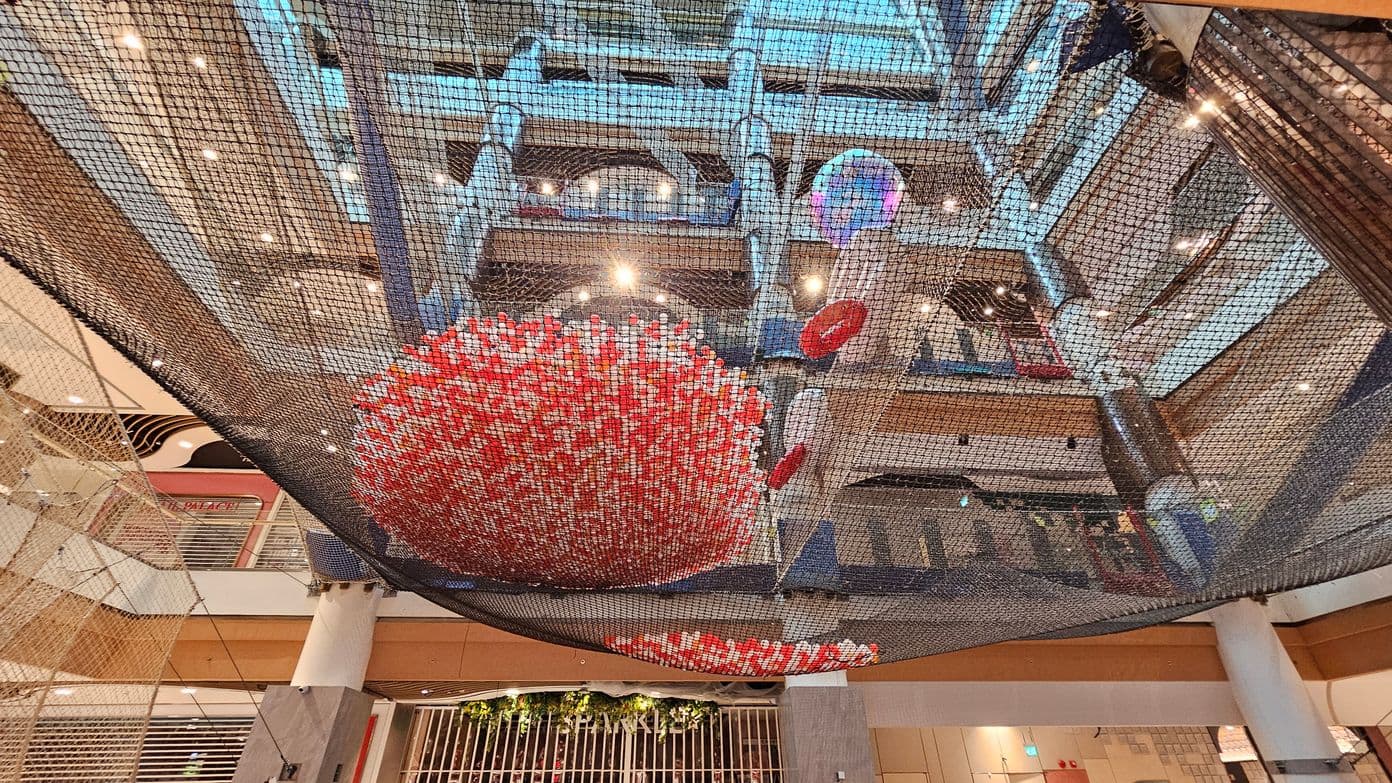 A large indoor space with a net structure suspended from the ceiling. In the centre, there's a spherical installation made of red and white elements. The background shows multiple floors with railings and a variety of lights. The ceiling is high with visible beams and spotlights. The floor is not visible, but there are columns supporting the structure. Some greenery is visible on the lower level.