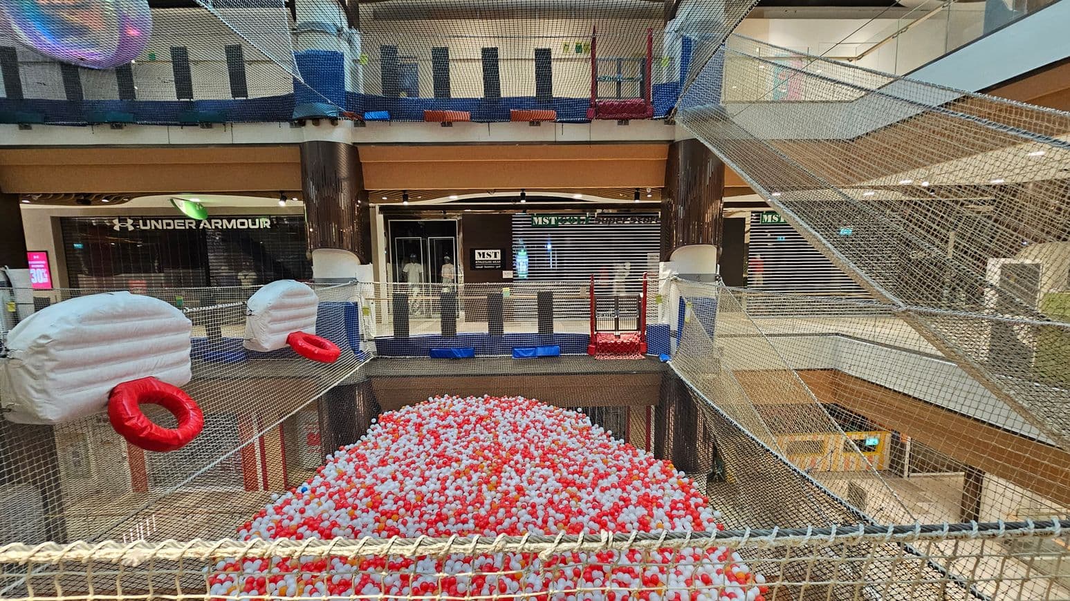 A shopping centre with a netted play area suspended above the ground. In the foreground, there are large white inflatable structures with red rings attached. Below, a pit filled with red and white balls. The background shows shopfronts, including Under Armour and MST, with closed shutters. The area is enclosed with netting, and there are multiple levels visible, with glass railings and a mix of wooden and metal elements.