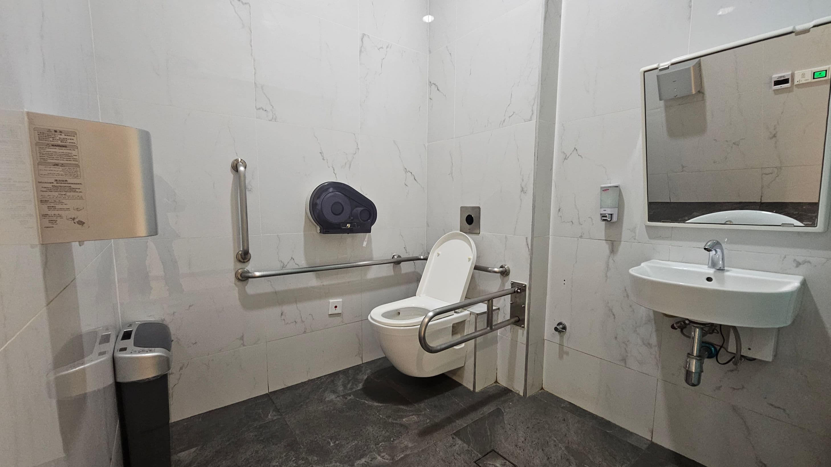 A public restroom with a white toilet featuring a raised seat and support rails on both sides. To the left, a waste bin and a wall-mounted instruction panel are visible. On the right, a white sink with a mirror above it and a soap dispenser to the side. The walls are covered in light-coloured tiles with a subtle marble pattern, and the floor is dark grey.