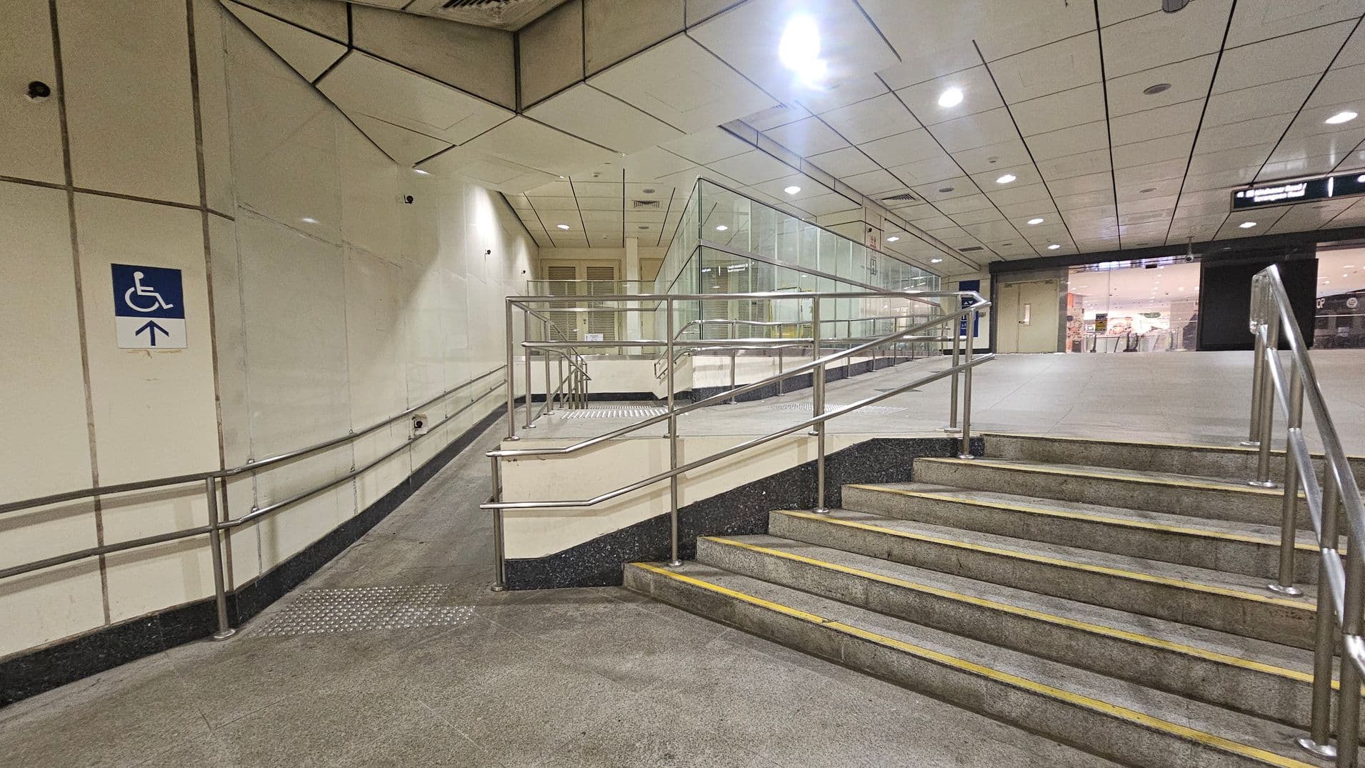 A public indoor space with a ramp and stairs. On the left, a ramp with metal railings leads upwards, marked by a blue sign indicating accessibility. The right side features a set of stairs with similar metal railings. The background shows a glass partition and a view into a well-lit area with shops. The ceiling is tiled with recessed lighting, and the floor is a mix of green and grey tiles.