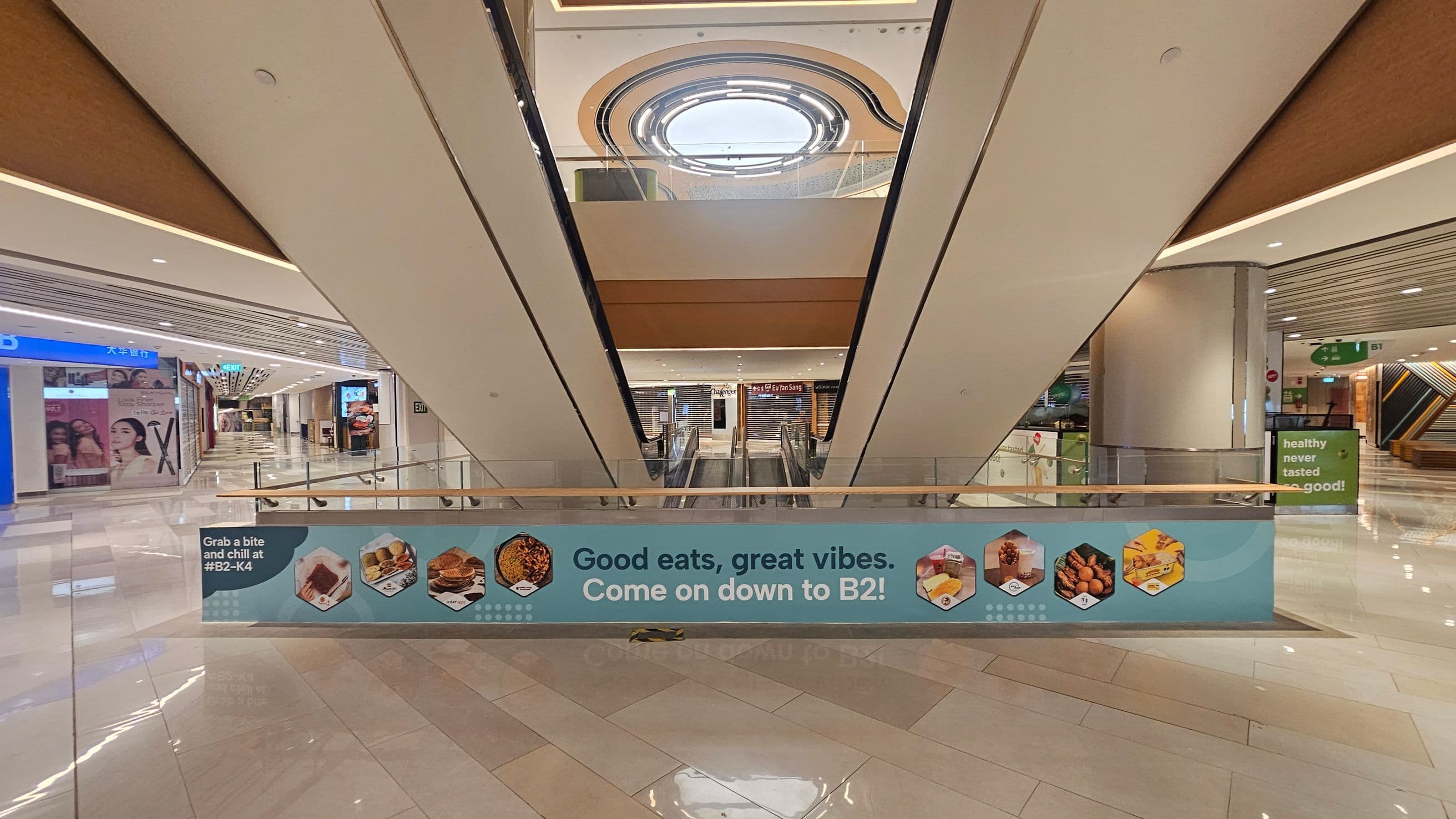 A shopping mall interior with a central escalator leading downwards. A blue banner across the escalator reads, "Good eats, great vibes. Come on down to B2!" with images of food items. The left side has shopfronts with advertisements, while the right side features a pillar and a sign saying "healthy never tasted so good!" The floor is shiny and tiled, and the ceiling has recessed lighting with a circular skylight above.