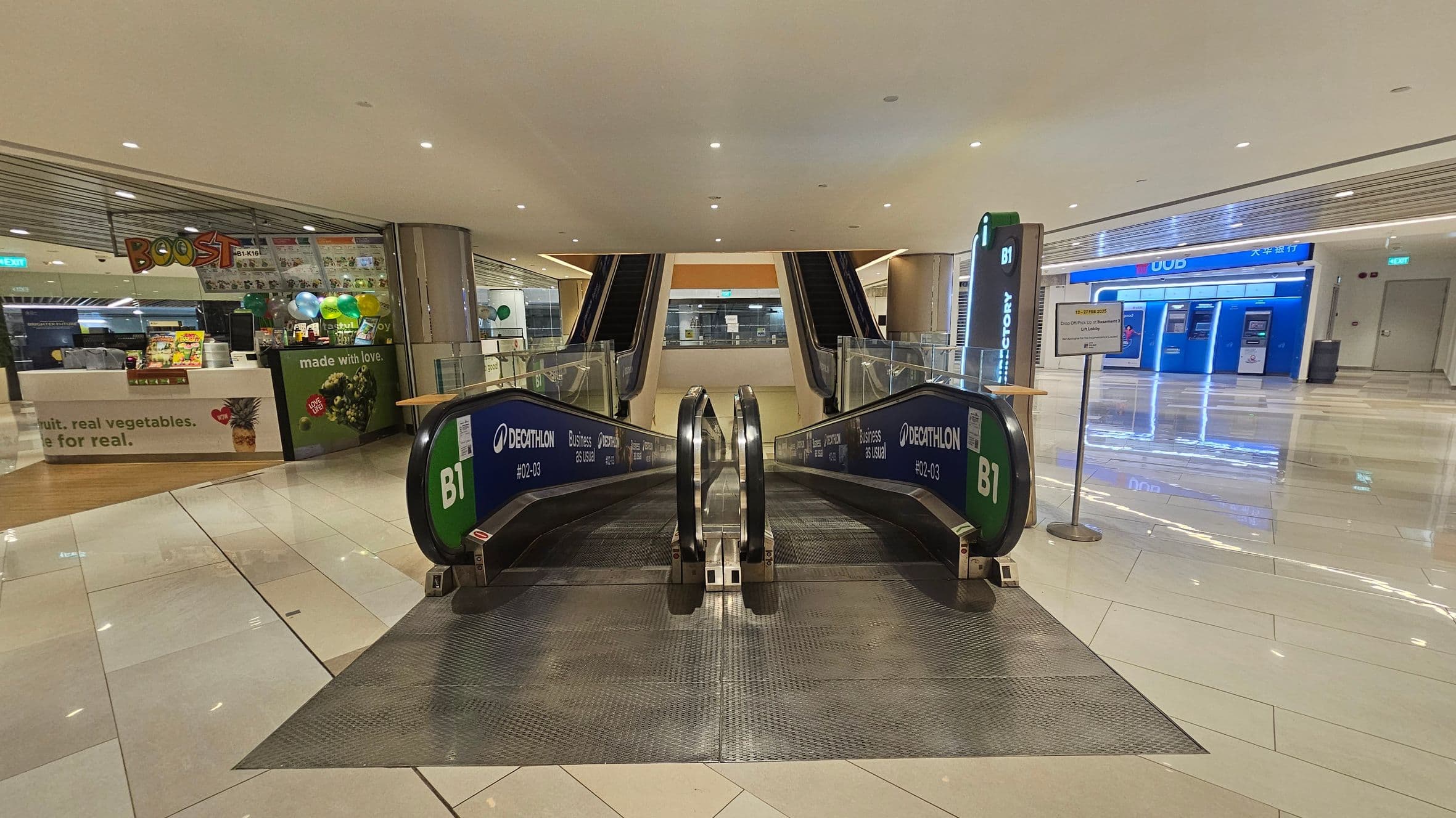 In the foreground, there are two escalators leading downwards, with green signage indicating "B1" and "Decathlon". On the left, there's a shop with a "Boost" sign, featuring a counter and colourful balloons. On the right, there is a bank area with several ATMs under a blue "UOB" sign. The floor is shiny and tiled, reflecting the ceiling lights. The ceiling is smooth with recessed lighting.