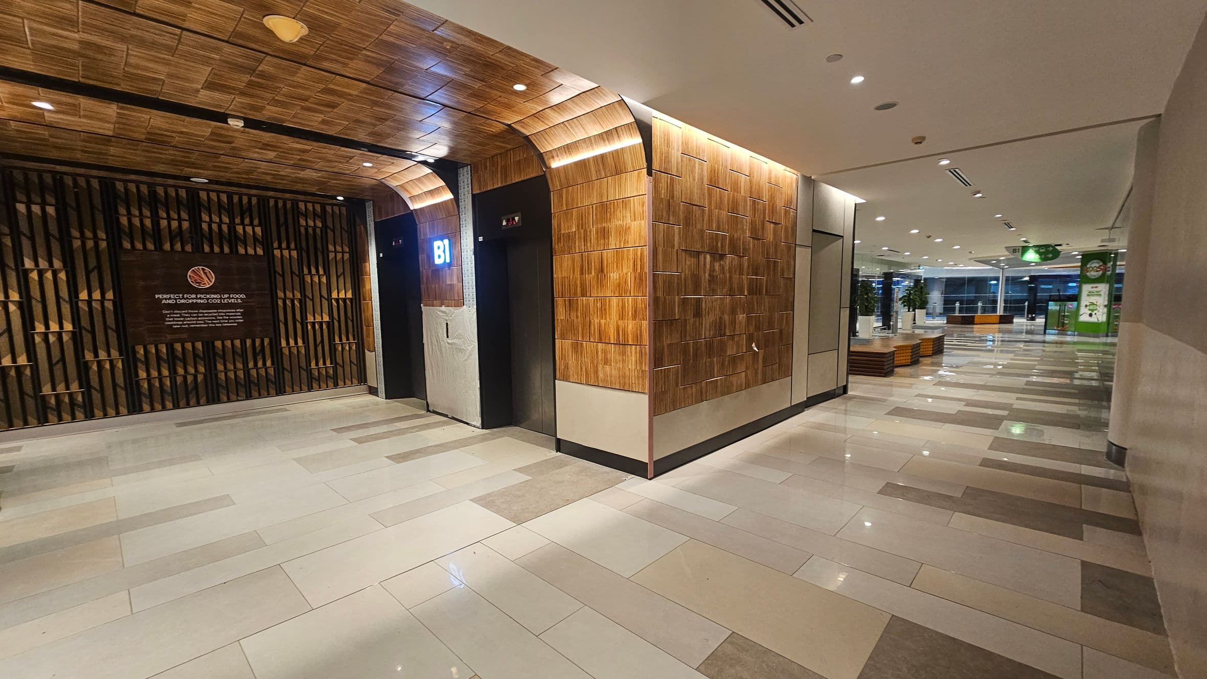 On the left, there is a wall with a wooden panel design and a sign with text. An elevator with a "B1" sign is next to it. The ceiling has a matching wooden pattern. On the right, the corridor extends with a tiled floor in shades of beige and grey. There are potted plants and seating areas along the corridor. The ceiling is white with recessed lighting. In the background, there are large windows and a green sign.