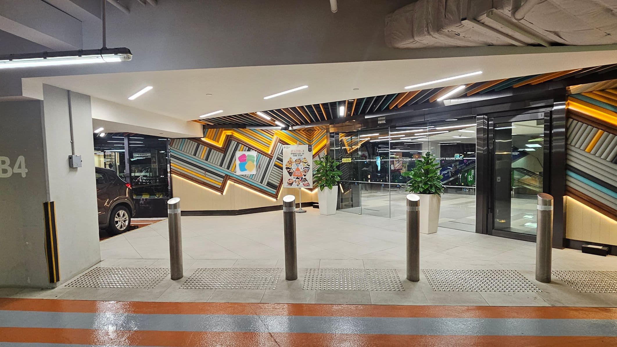 A modern car park entrance with a geometric, colourful wall design featuring diagonal lines in shades of orange, blue, and grey. On the left, a black car is partially visible next to a grey pillar marked "B4". In the centre, there are three metallic bollards and a sign with images of food. Two potted plants are placed near the glass doors on the right, leading to an interior space. The ceiling has exposed pipes and bright lighting. The floor is tiled with a textured strip near the bollards.