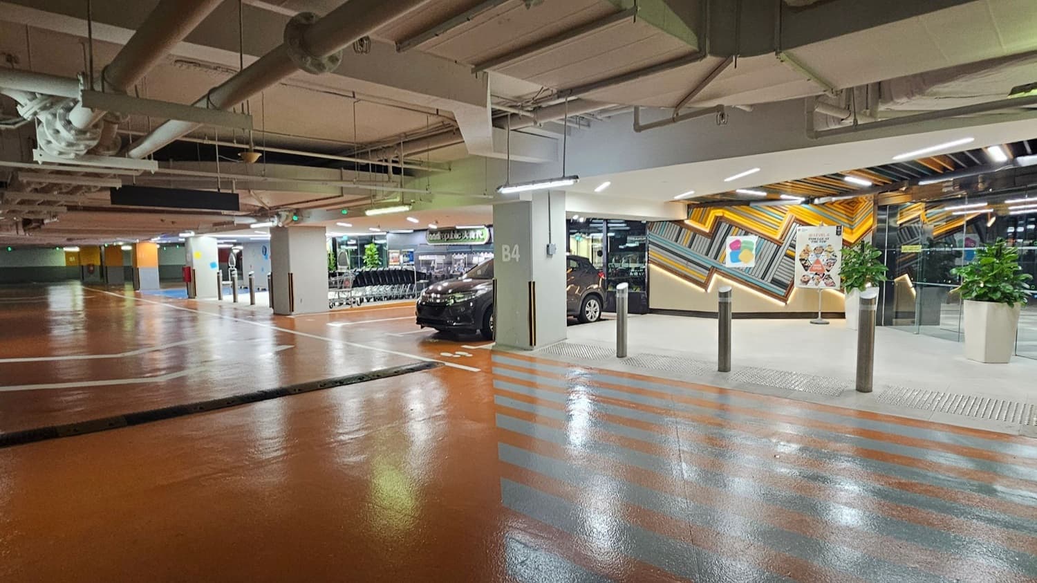 A brightly lit underground car park with a shiny, polished floor. On the left, there are empty parking spaces and a row of shopping trolleys. In the centre, a black car is parked next to a pillar marked "B4". On the right, a colourful geometric wall design with a poster and two large potted plants. The ceiling is exposed with visible pipes and lighting fixtures.