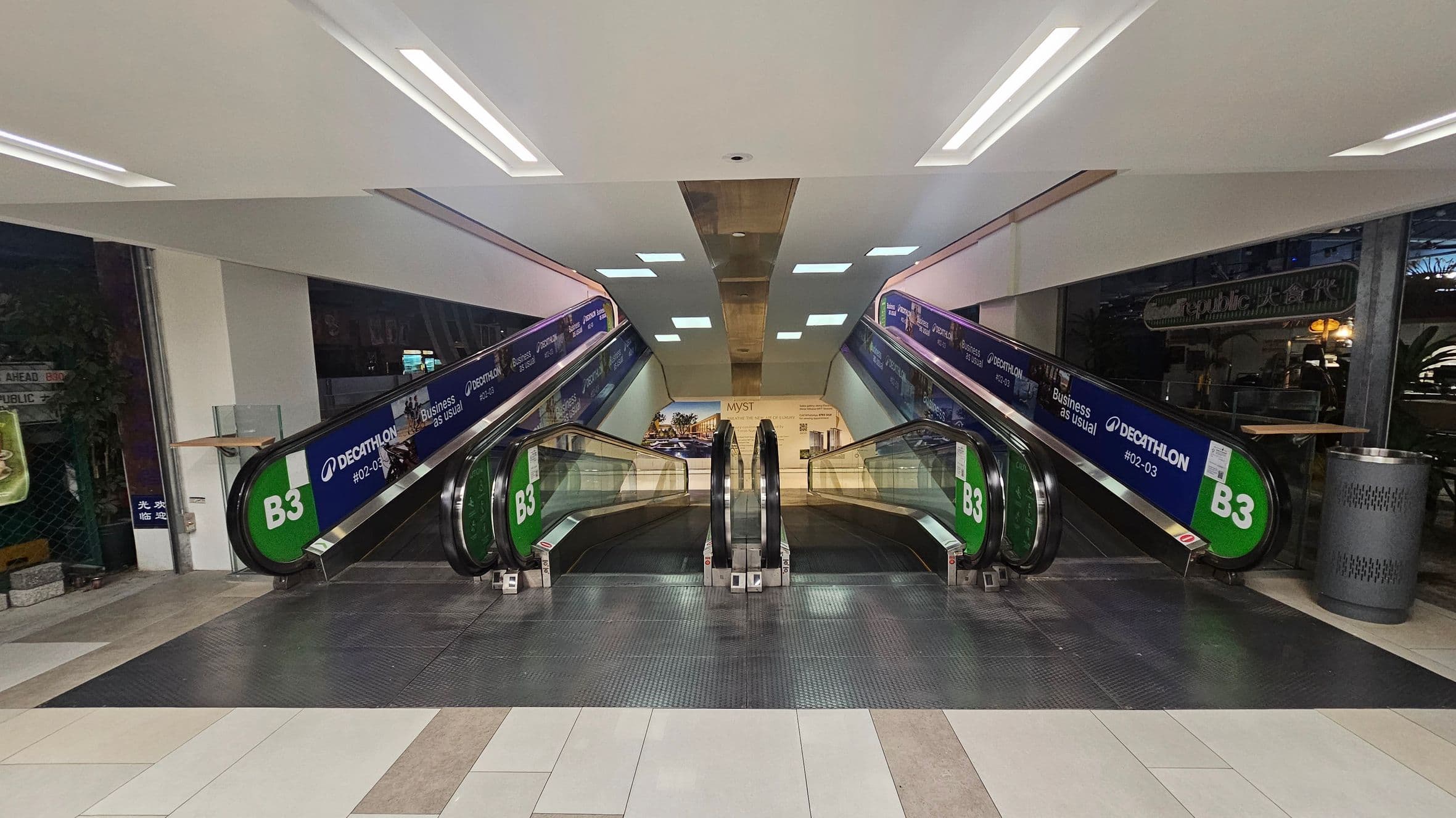 Two parallel escalators in a shopping centre, with green signs marked "B3" on the sides. The left escalator is for descending, and the right is for ascending. Advertisements for a store are visible along the sides. The floor is tiled in a mix of light and dark shades, and the ceiling is white with recessed lighting. A trash bin is on the right side near the escalator. In the background, there is a large poster with images and text.