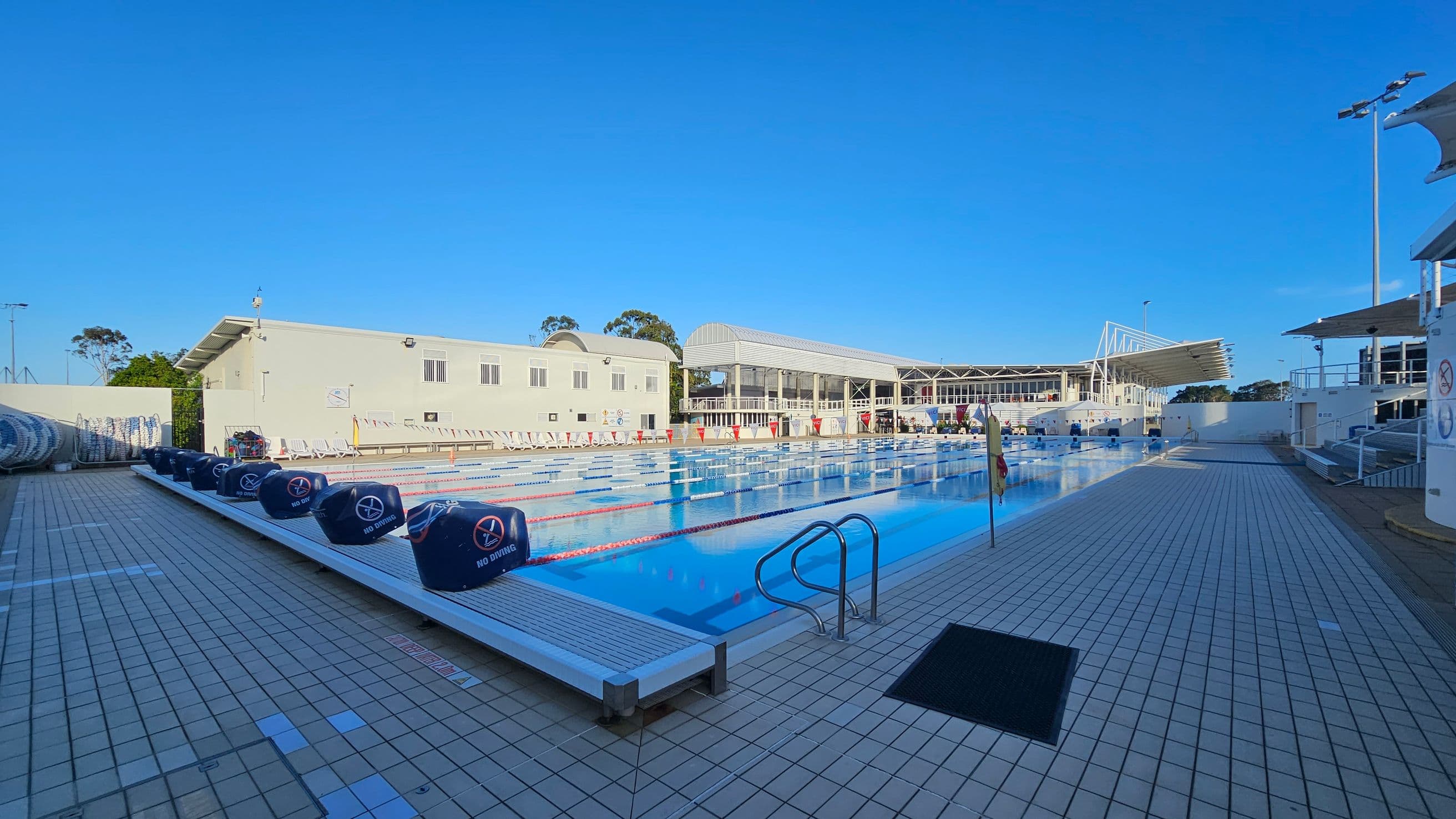 An outdoor swimming pool with multiple lanes, surrounded by a tiled deck. On the left, there are blue lane dividers and a building with windows. On the right, a set of metal pool ladders and a covered seating area are visible. In the background, a large structure with a white roof and open sides is present. The pool is calm, and the area is well-lit with tall lights.