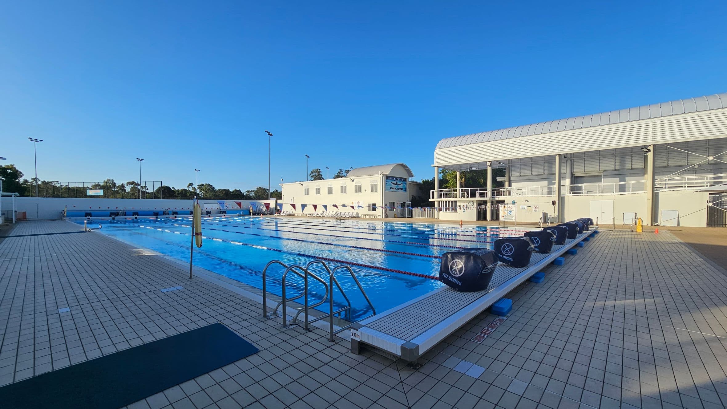 An outdoor swimming pool with clear blue water and lane dividers stretches across the foreground. On the right, a modern building with large windows and a curved roof stands adjacent to the pool. On the left, a smaller building with a similar roof is visible. The pool deck is tiled, and there are metal ladders for entry. In the background, trees and tall floodlights are visible. The sky is clear and blue.