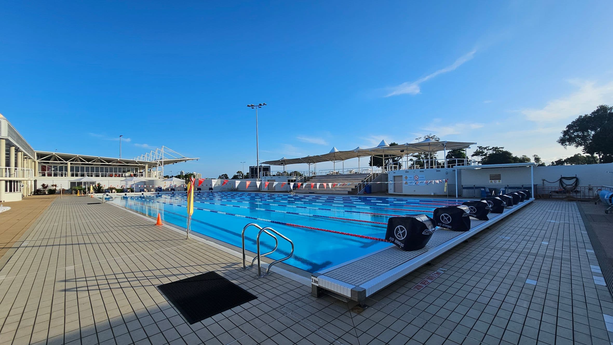 An outdoor swimming pool with clear blue water and lane dividers. On the left, a modern building with large windows and a sloped roof. The pool deck is tiled with a mix of light and dark squares. On the right, a row of covered seating areas with shade structures. Several "No Diving" signs are visible along the pool's edge. In the background, trees and a clear sky. A few orange cones are placed near the poolside.