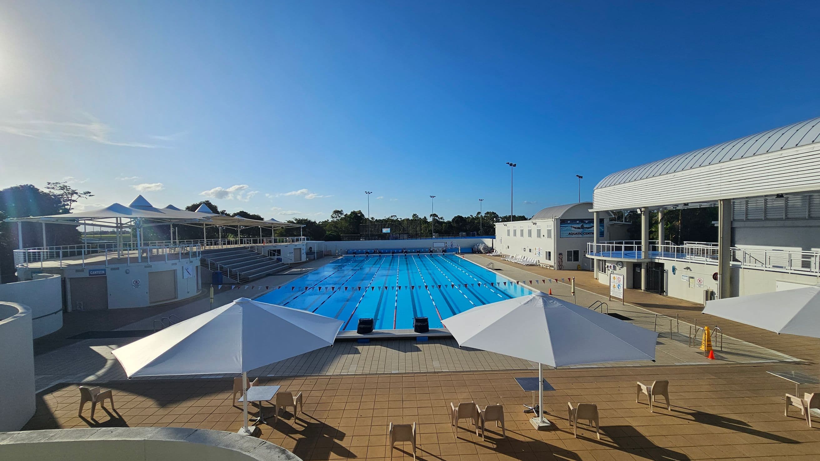 An outdoor swimming pool with clear blue water is in the centre, surrounded by a tiled deck. On the left, there are shaded seating areas with white canopies and chairs. On the right, a building with a curved roof and multiple levels overlooks the pool. In the background, trees and a clear sky are visible. The foreground features more seating and umbrellas, with a few scattered chairs and tables.