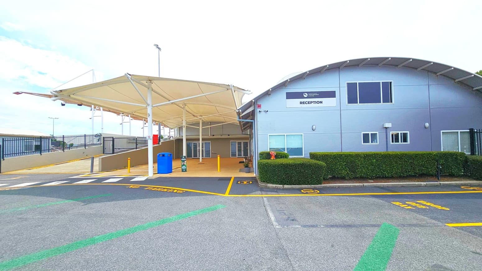 A building with a curved roof on the right, featuring a sign that reads "RECEPTION". The entrance is covered by a large, white canopy supported by metal poles. A blue bin and a small sign are near the entrance. The foreground has a marked pedestrian crossing and yellow lines on the asphalt. A hedge runs along the building's side, and a black metal fence is visible on the left. The area is well-maintained and organised.
