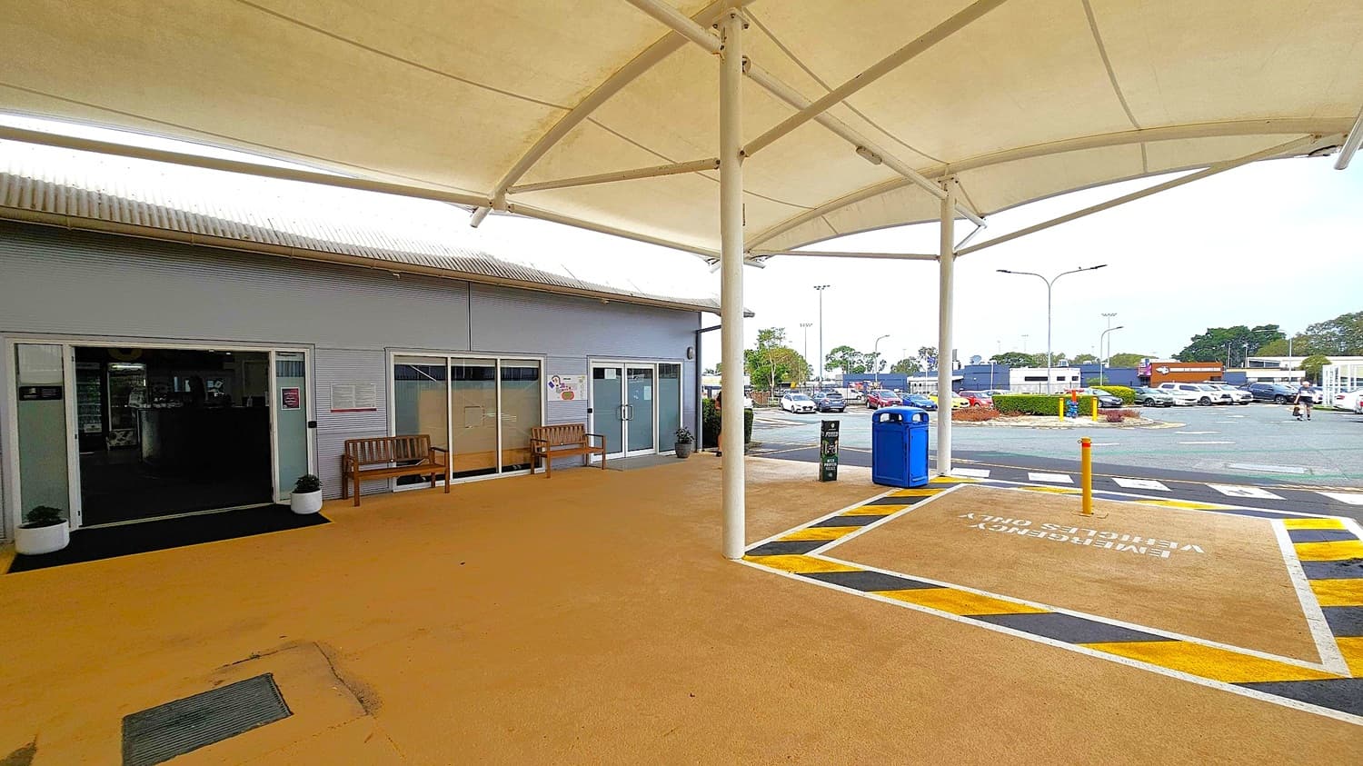 A covered outdoor area with a beige floor and a white canopy ceiling. On the left, a building with grey walls and large glass doors and windows, flanked by wooden benches and potted plants. In the centre, a white pole supports the canopy. On the right, a blue bin and a signpost are near a marked emergency vehicle parking area. In the background, a car park with several vehicles and trees is visible.