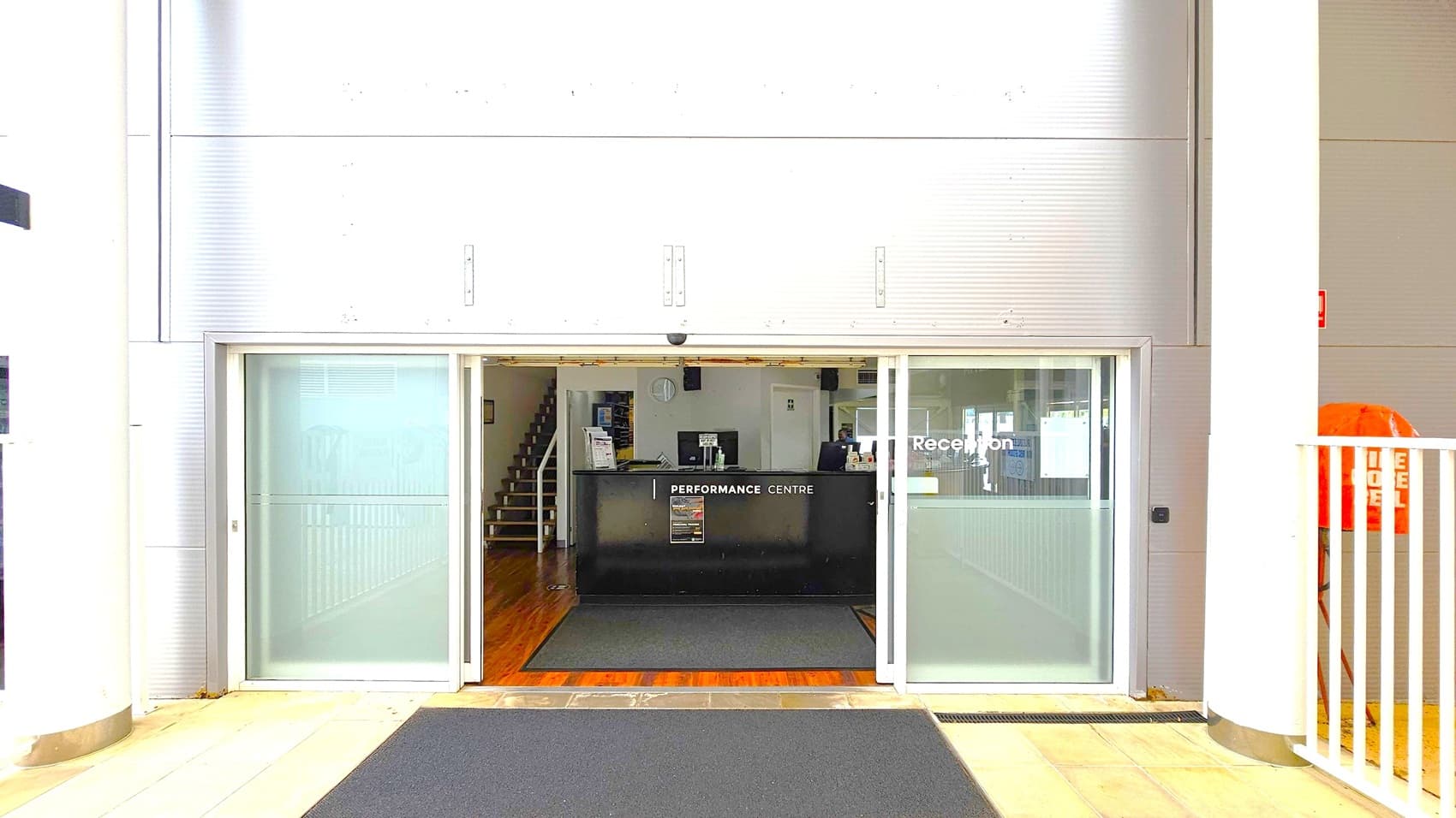 A reception area with a black desk labelled "Performance Centre" in the middle. The entrance has sliding glass doors with "Reception" written on the right door. Inside, a staircase is visible to the left. The floor is tiled with a dark mat leading to the entrance. The walls are metallic with vertical lines, and there are large white columns on either side. An orange object is partially visible on the right, behind a white railing.