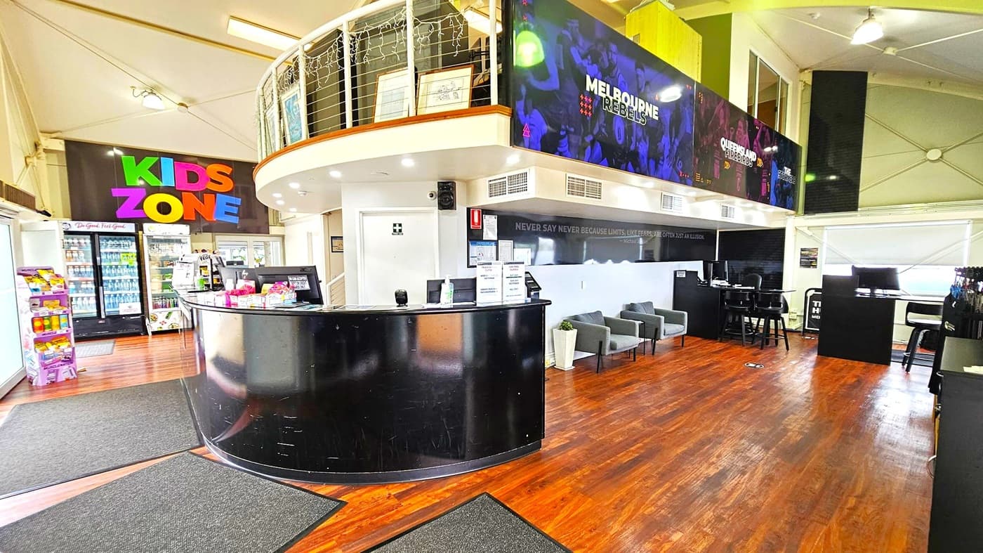 A spacious interior with a wooden floor and a high ceiling. On the left, there's a colourful "KIDS ZONE" sign above a refrigerated drinks display and snack shelves. In the centre, a curved black reception desk is adorned with various items. To the right, a seating area with grey chairs and a high table with stools. Above, a mezzanine level with railings and framed pictures. The walls feature large banners with sports team names. Ceiling lights illuminate the space.