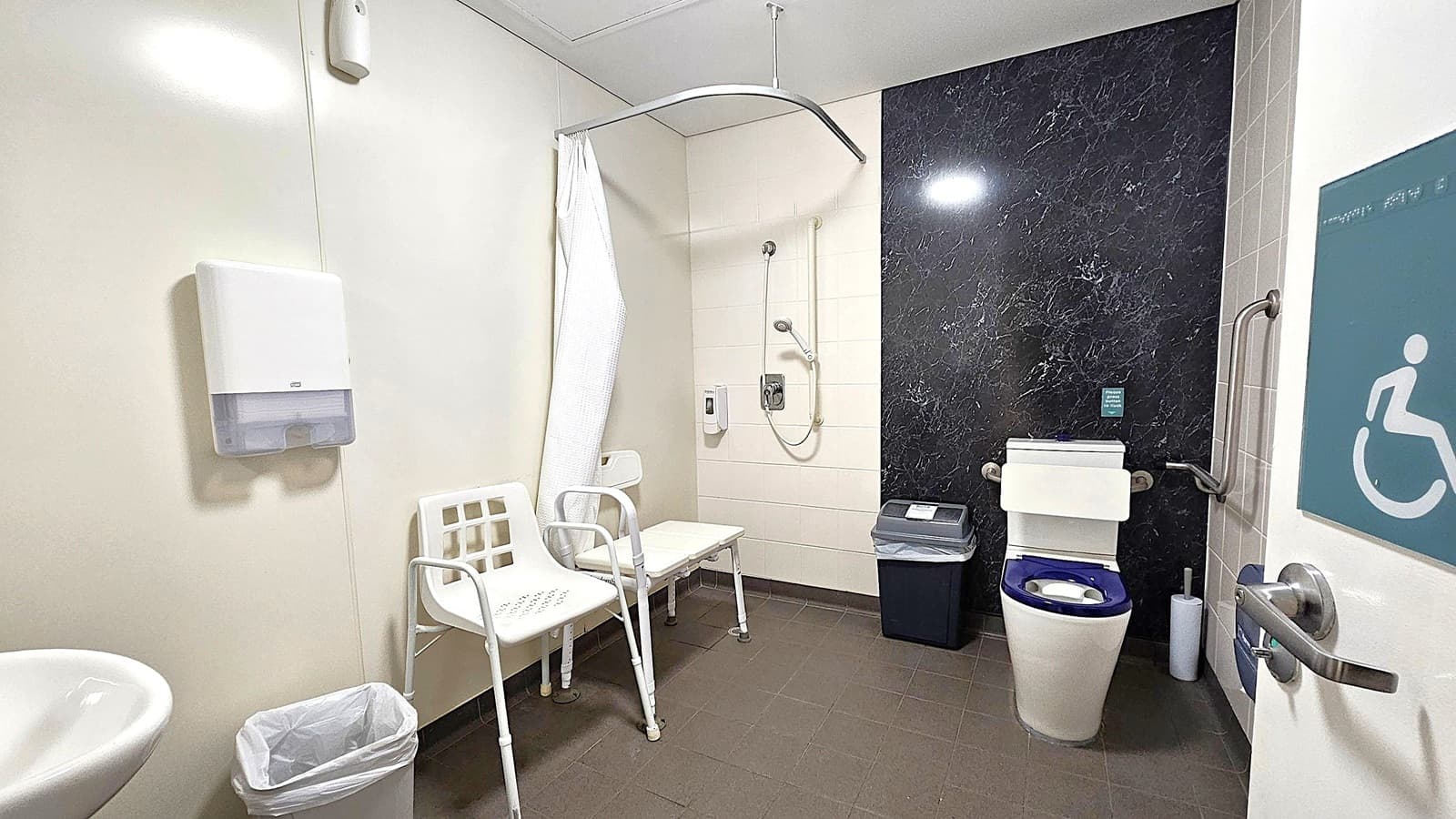 Accessible bathroom with a white sink and paper towel dispenser on the left wall. A waste bin is below. In the centre, a shower area with a curtain and a white plastic chair. The right side has a black marble wall with a toilet featuring a backrest and grab bars. A bin is nearby. The floor is dark tiled, and the ceiling is plain white. A door with a disability sign is partially open on the right.