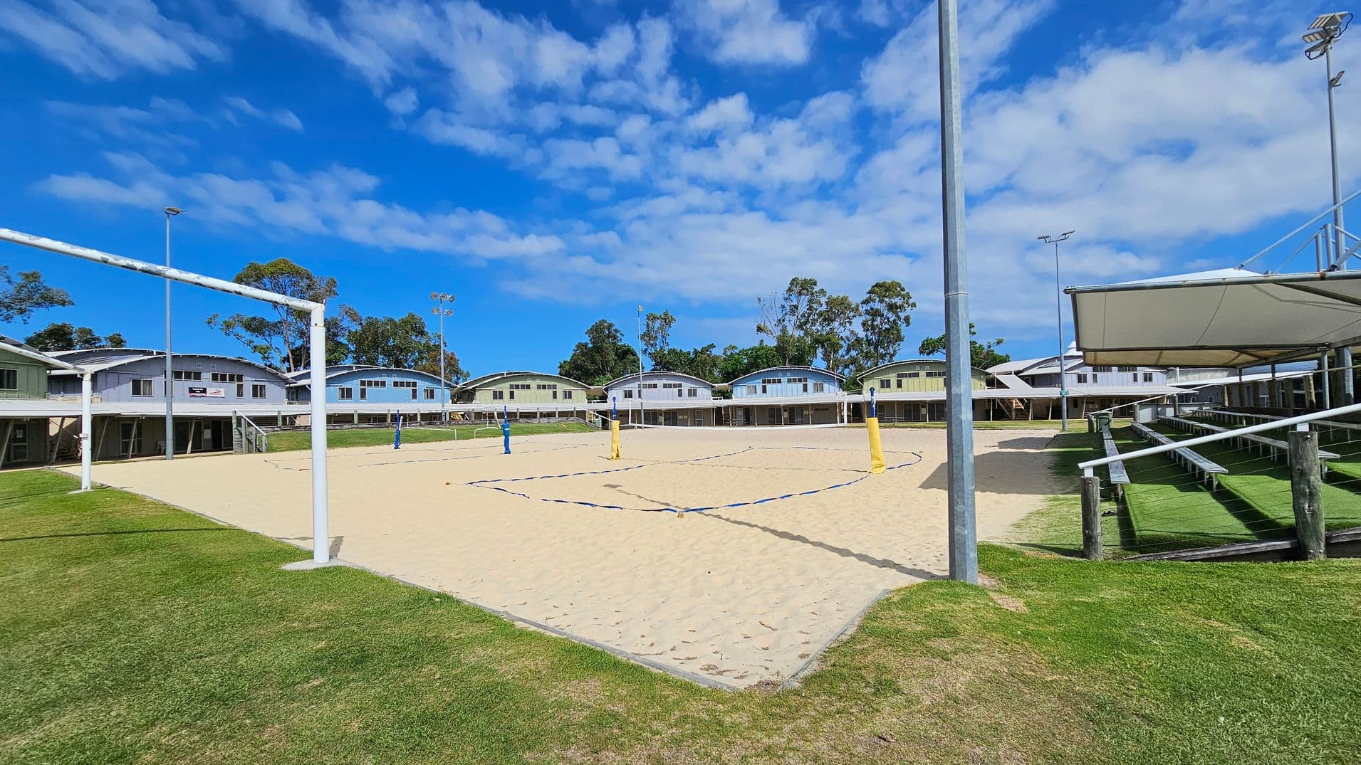 On the left, grass borders a sandy volleyball court framed by white poles and blue boundary lines. In the middle, the sandy court features yellow and blue net posts, with buildings in pale green, cream, and white in the background, all with curved roofs and supported by stilts. On the right, a covered seating area with tiered white benches overlooks the court, resting on artificial green grass. Floodlights on tall poles are positioned around the area.