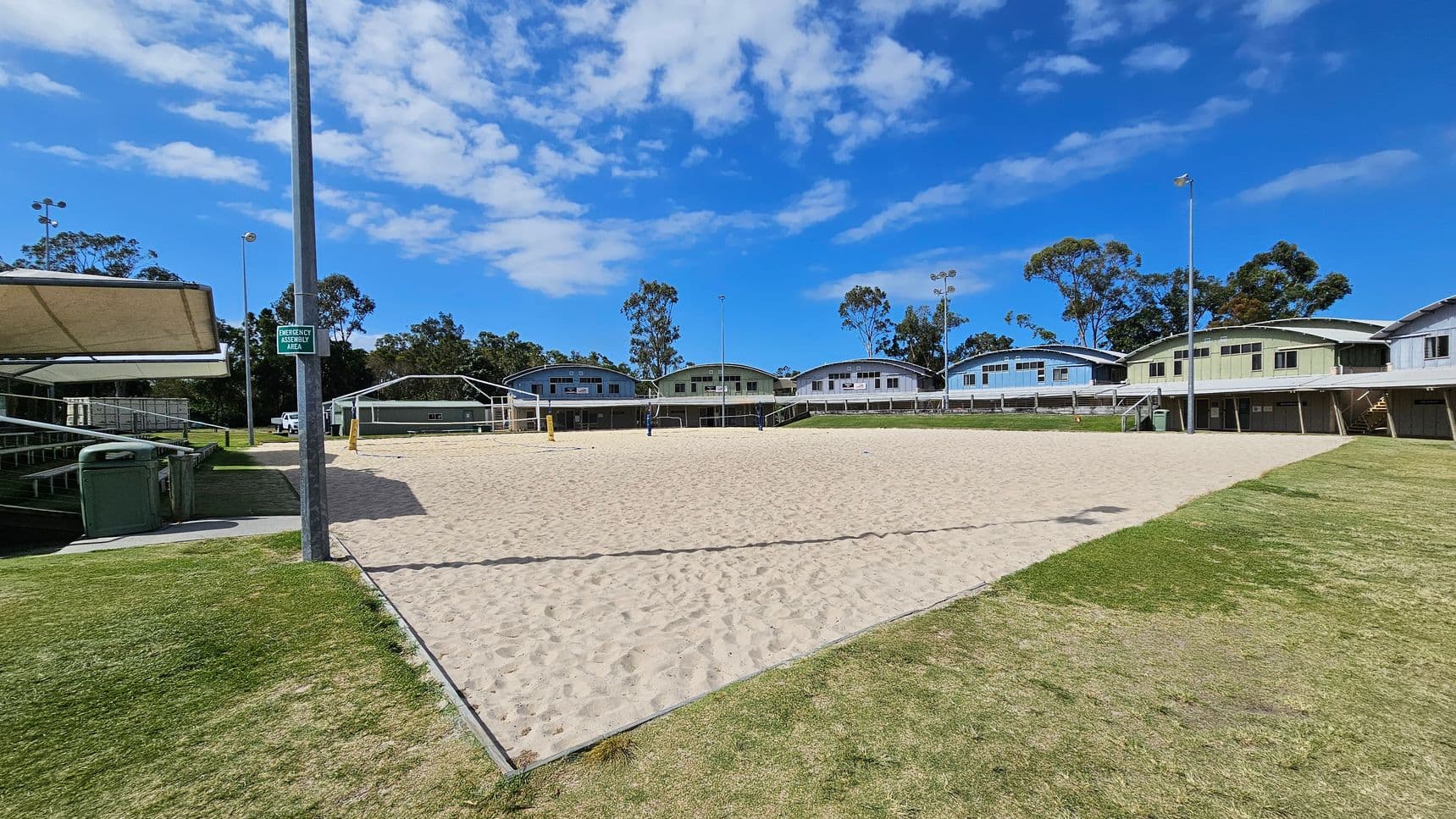 On the left, tiered metal seating is covered by a beige shade structure, next to two green bins on a concrete path that borders a grassy area. In the middle, a sand volleyball court has blue and yellow net posts, surrounded by short grass. In the background, pale green and blue arched-roof buildings are evenly spaced alongside tall trees. On the right, grassy areas extend to another shade structure and more seating with stair access. Floodlights are positioned around the space.