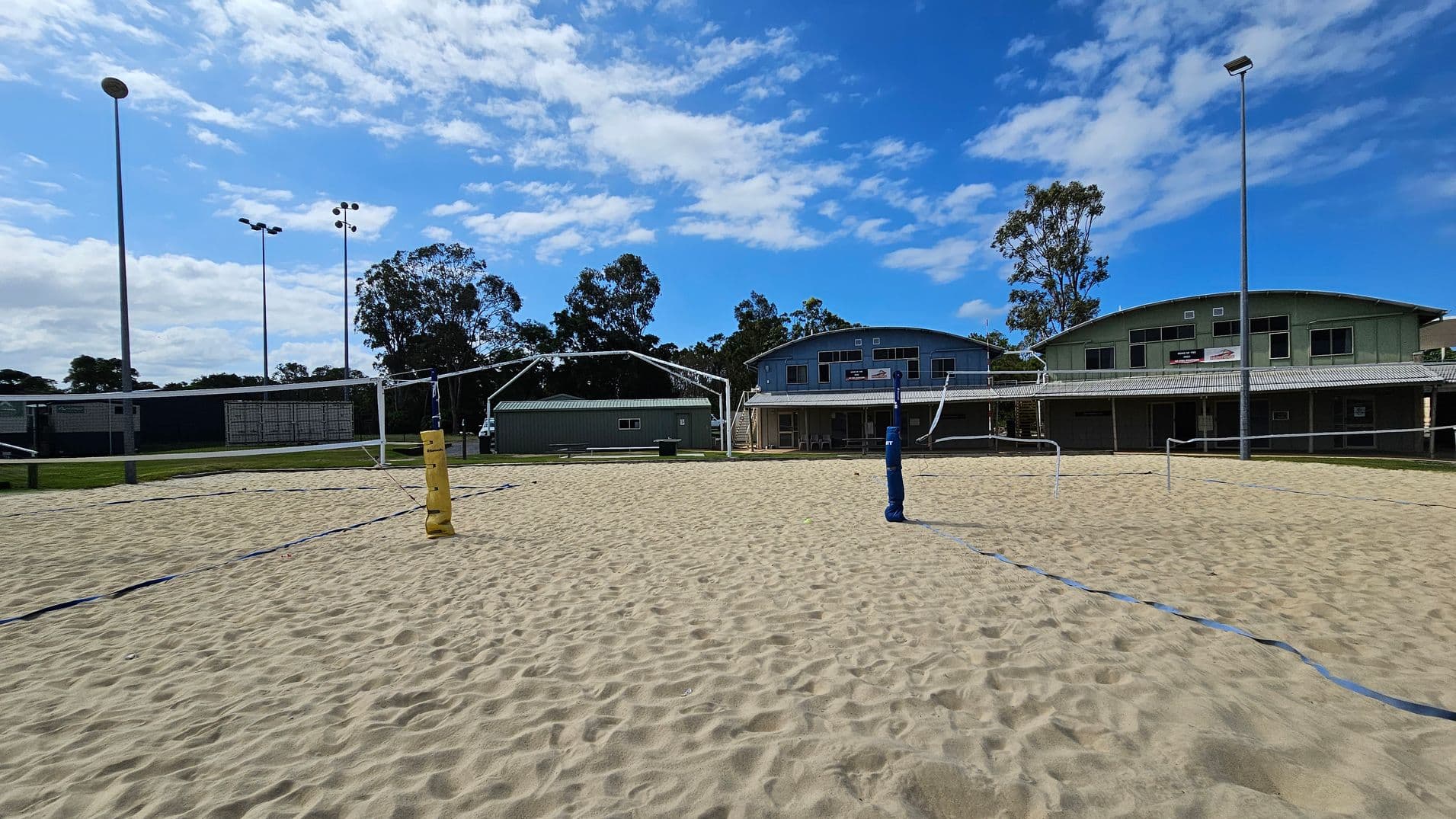 On the left, a shaded seating area with tiered metal benches sits beside a concrete path and grassy ground. In the middle, a large sandy volleyball court with blue boundary lines and yellow posts extends across the space. On the right, the sandy court is bordered by grass leading to pale green and blue arched-roof buildings with support stilts. Tall floodlights are positioned across the area, and the ground transitions between sand, grass, and pavement.