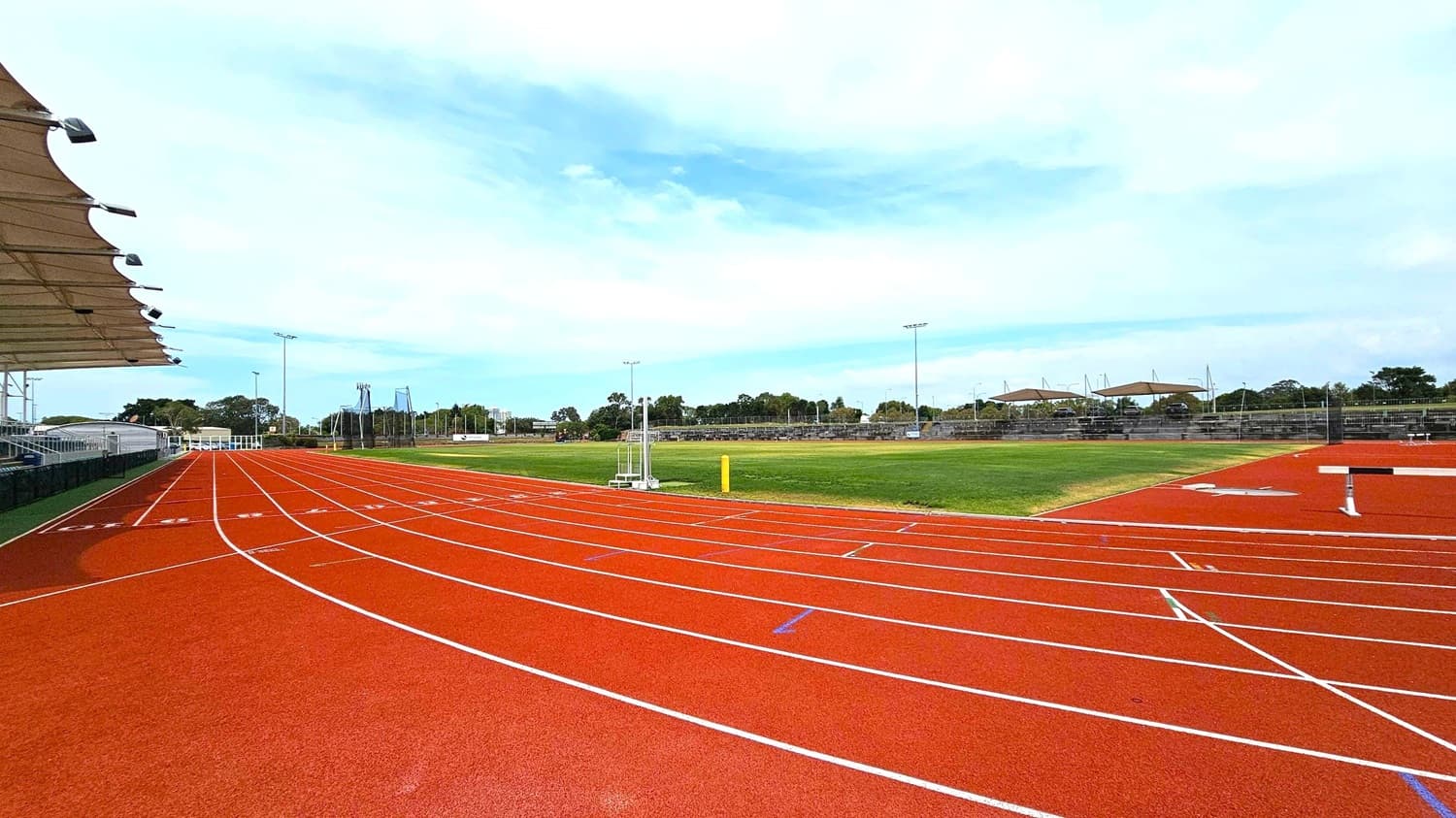 A red athletics track with white lane markings curves to the left. On the left side, there is a covered seating area with a canopy and lights. To the right, a green grass field is visible. In the background, there are more seating areas with shade structures and some trees. Floodlights are positioned around the track and field. The sky is overcast.