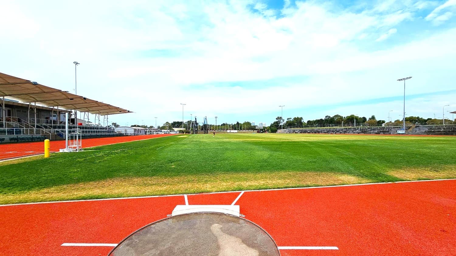 A sports field with a red running track encircling a large grassy area. In the foreground, a shot put circle is visible. On the left, there is a covered seating area with a canopy and metal bleachers. The right side features more bleachers and a few floodlights. In the background, there are trees and additional sports equipment, including goalposts and nets. The sky is overcast.