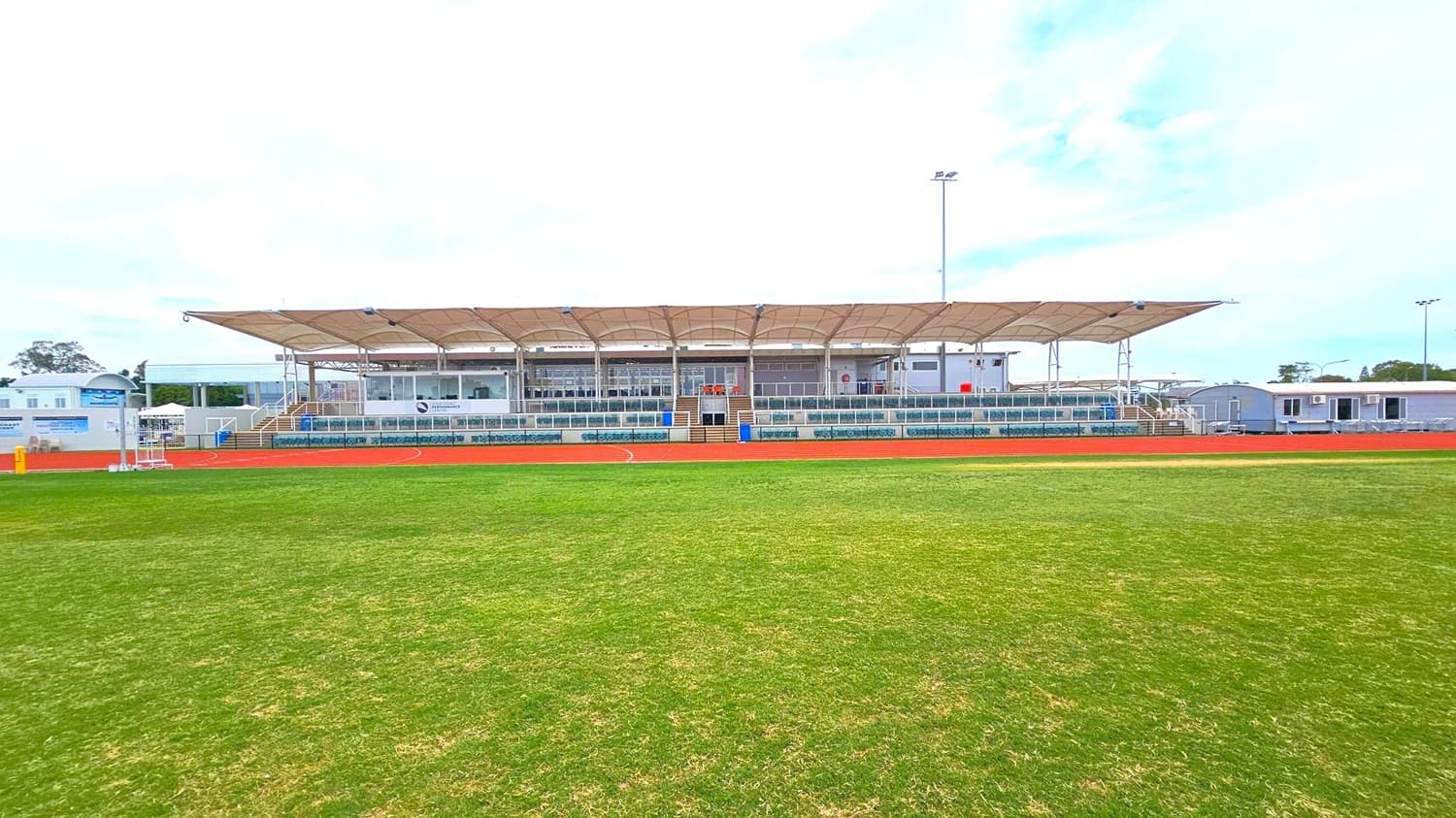 A sports stadium with a large, covered grandstand in the centre. The grandstand has a beige canopy roof and multiple rows of seating. In front of the grandstand is a red athletics track, and beyond that, a green grass field. On either side of the grandstand, there are smaller structures and additional seating areas. Tall floodlights are visible above the grandstand. The sky is overcast.
