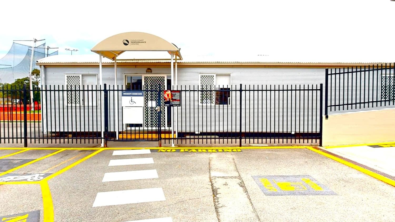 A single-storey building with a corrugated metal exterior and barred windows is behind a black metal fence. The entrance has a small awning with a sign above it. In front of the building, there is a marked pedestrian crossing and a "No Parking" area painted on the ground. The pavement is grey with yellow lines, and there is a designated disabled parking space. To the left, there are netted structures and some trees in the background.