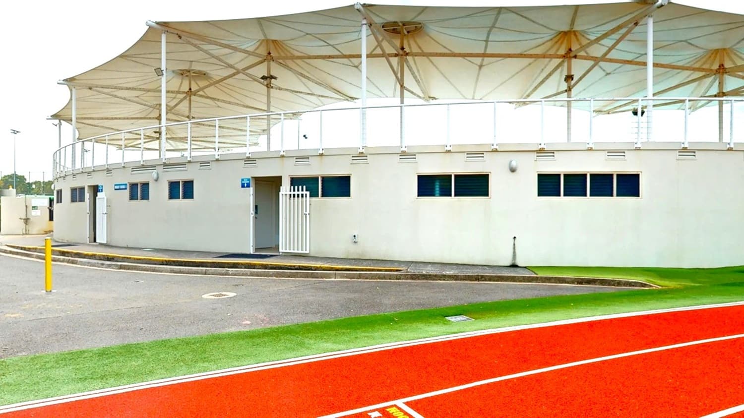 A building with a white exterior and small rectangular windows is in the centre. It has a large, white, tensile fabric roof supported by poles. In front of the building, there is a red running track with white lines. The ground is a mix of grey pavement and green grass. A yellow bollard is visible on the left side near the pavement. The area appears to be part of a sports facility.