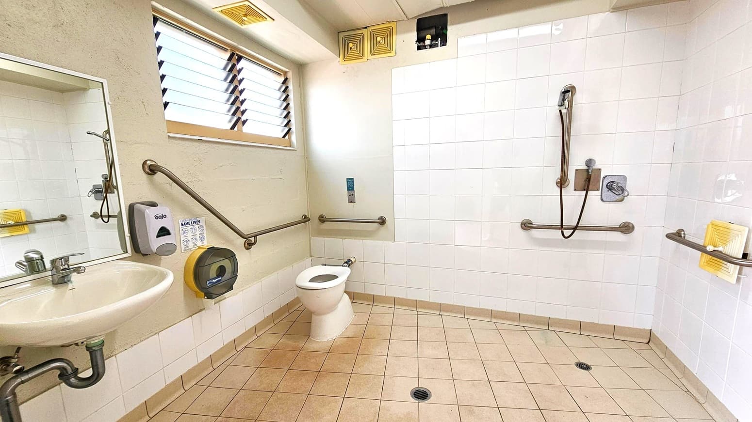 A bathroom with beige tiled flooring and white tiled walls. On the left, a sink with a mirror above it, a soap dispenser, and a toilet paper holder. A toilet is positioned centrally, with grab bars around it. On the right, a shower area with a handheld showerhead and additional grab bars. A small window with louvres is above the sink, and there are ventilation grilles near the ceiling. A drain is visible on the floor.