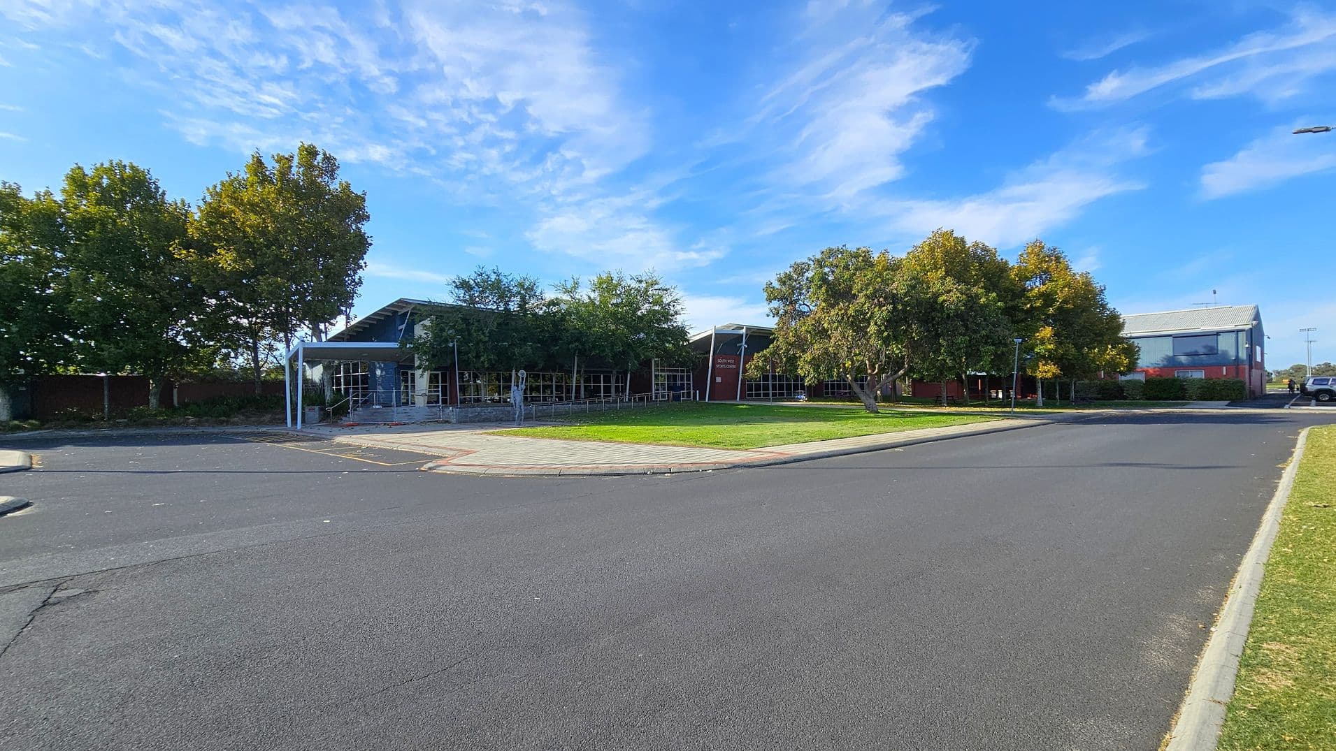 A wide road curves around a grassy area in the foreground. On the left, there are tall trees with dense foliage. In the centre, a modern building with large windows and a slanted roof is partially obscured by smaller trees. To the right, another building with a grey roof and red brick walls is visible. A few cars are parked near the buildings. The sky is clear with some wispy clouds.