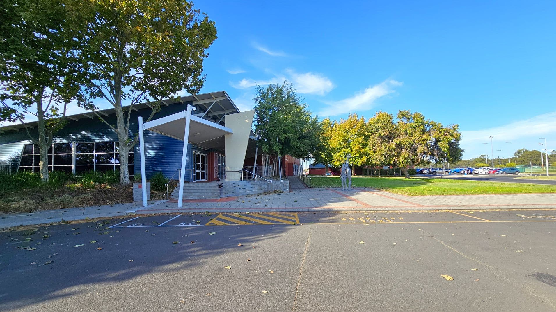 A modern building with a slanted roof and large windows is on the left, partially shaded by tall trees. The entrance features a white canopy and steps leading up to it. On the right, a paved area with a sculpture of a person and more trees in the background. A car park with several vehicles is visible further right. The ground is a mix of asphalt and paving, with some fallen leaves scattered around.