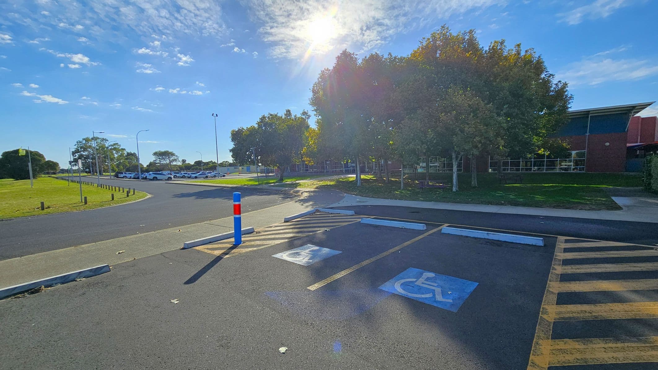 A parking area with two marked disabled parking spaces in the foreground. The ground is asphalt with painted symbols. A blue and red bollard is near the spaces. In the background, there are trees casting shadows and a building with large windows. To the left, a grassy area with a row of parked cars along a road. The sun is shining brightly, creating lens flare effects.
