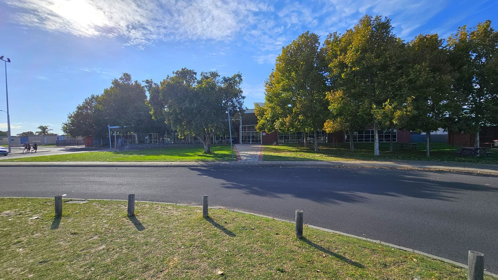 A paved road runs horizontally across the foreground with a grassy verge and wooden posts. In the middle, a path leads through a row of trees with green and yellow leaves, towards a building with large windows. On the left, a few people walk near a lamppost, and a car is parked nearby. The background features more trees and a clear sky.