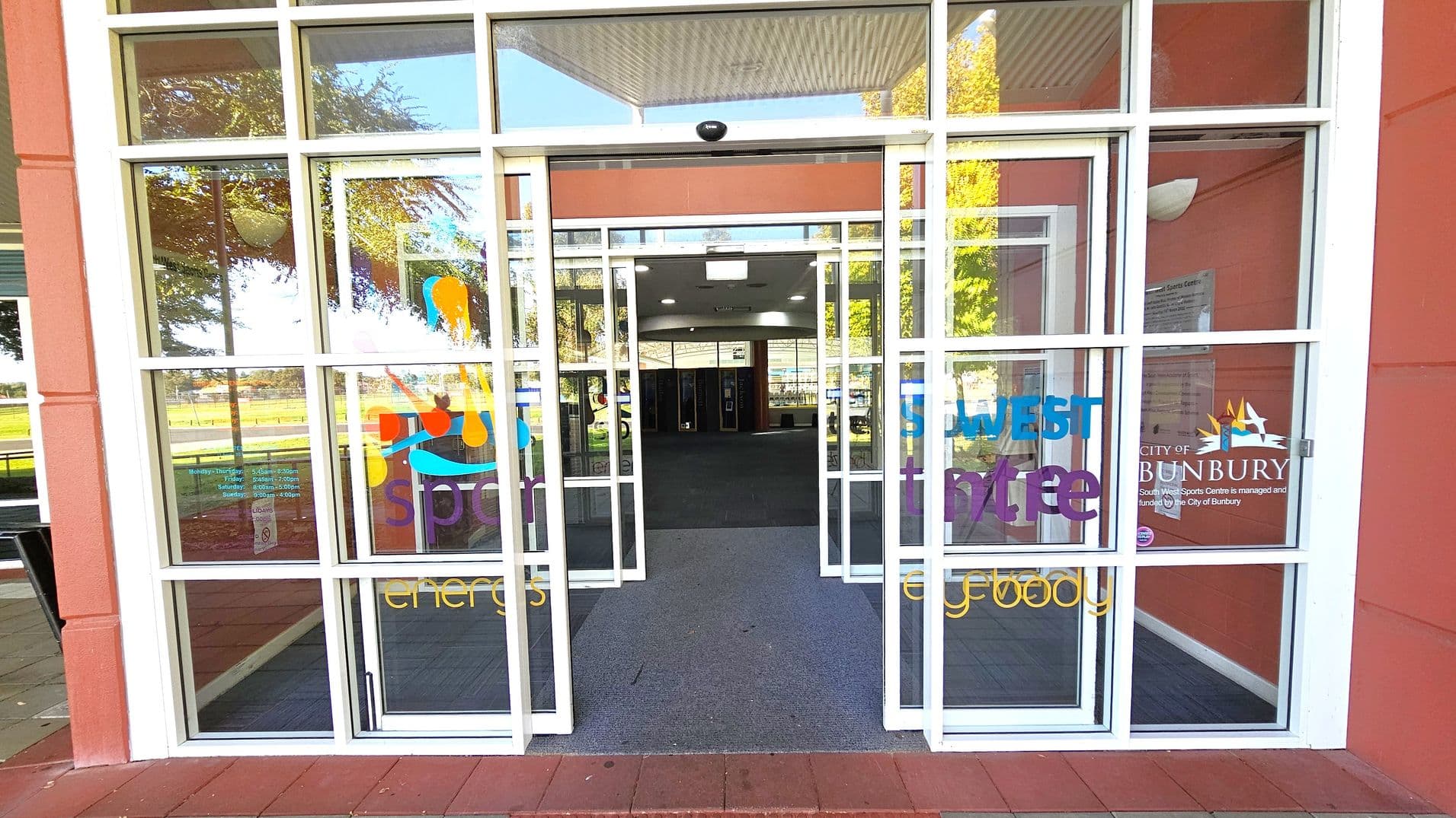 Glass entrance doors with white frames, featuring colourful text and graphics. The words "South West Sports Centre" and "City of Bunbury" are visible. Inside, a carpeted floor leads to a spacious interior with ceiling lights. Reflections of trees and outdoor seating are visible on the glass. The exterior walls are a reddish-brown colour.