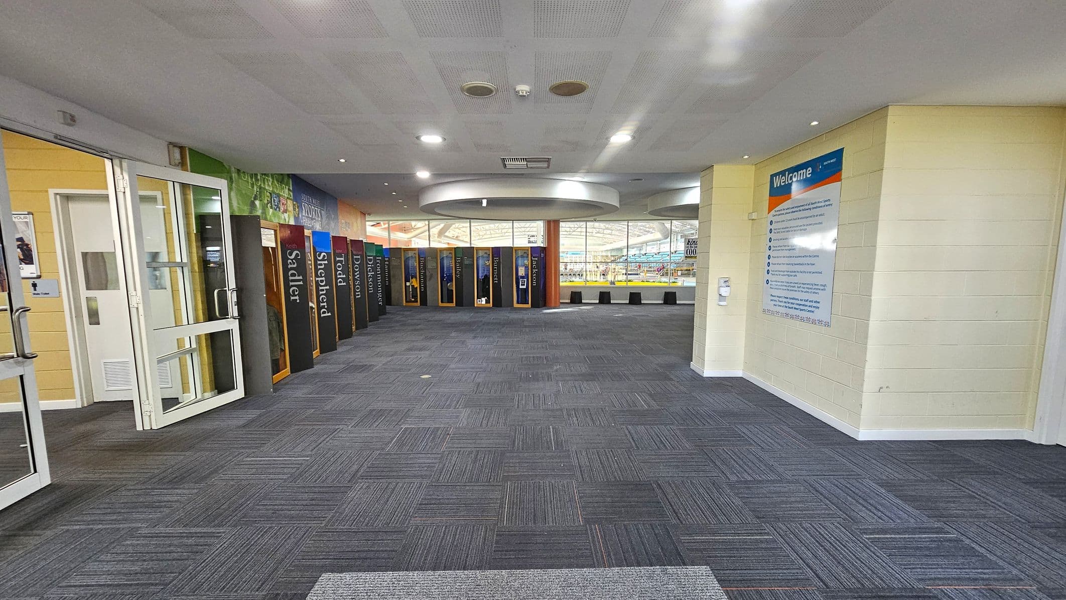 A spacious indoor area with a grey carpeted floor. On the left, there are glass doors and a sign indicating restrooms. The right side features a large welcome sign on a light-coloured wall. In the centre, a series of colourful lockers line the wall, each with a name. The ceiling is white with recessed lighting and circular fixtures. In the background, large windows reveal a glimpse of an indoor pool area.