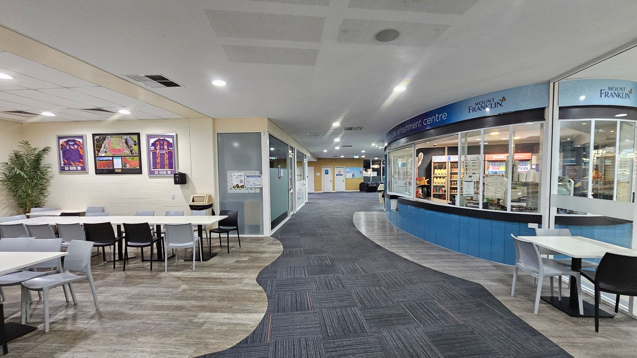 On the left, there are white tables with a mix of black and grey chairs, and framed pictures on the wall above. A potted plant is in the corner. The floor has a mix of green and brown tiles. On the right, there's a refreshment centre with a curved counter and glass display, featuring various drinks and snacks. The floor transitions to grey tiles. The ceiling is white with recessed lighting. In the background, there are doors and a hallway leading further inside.