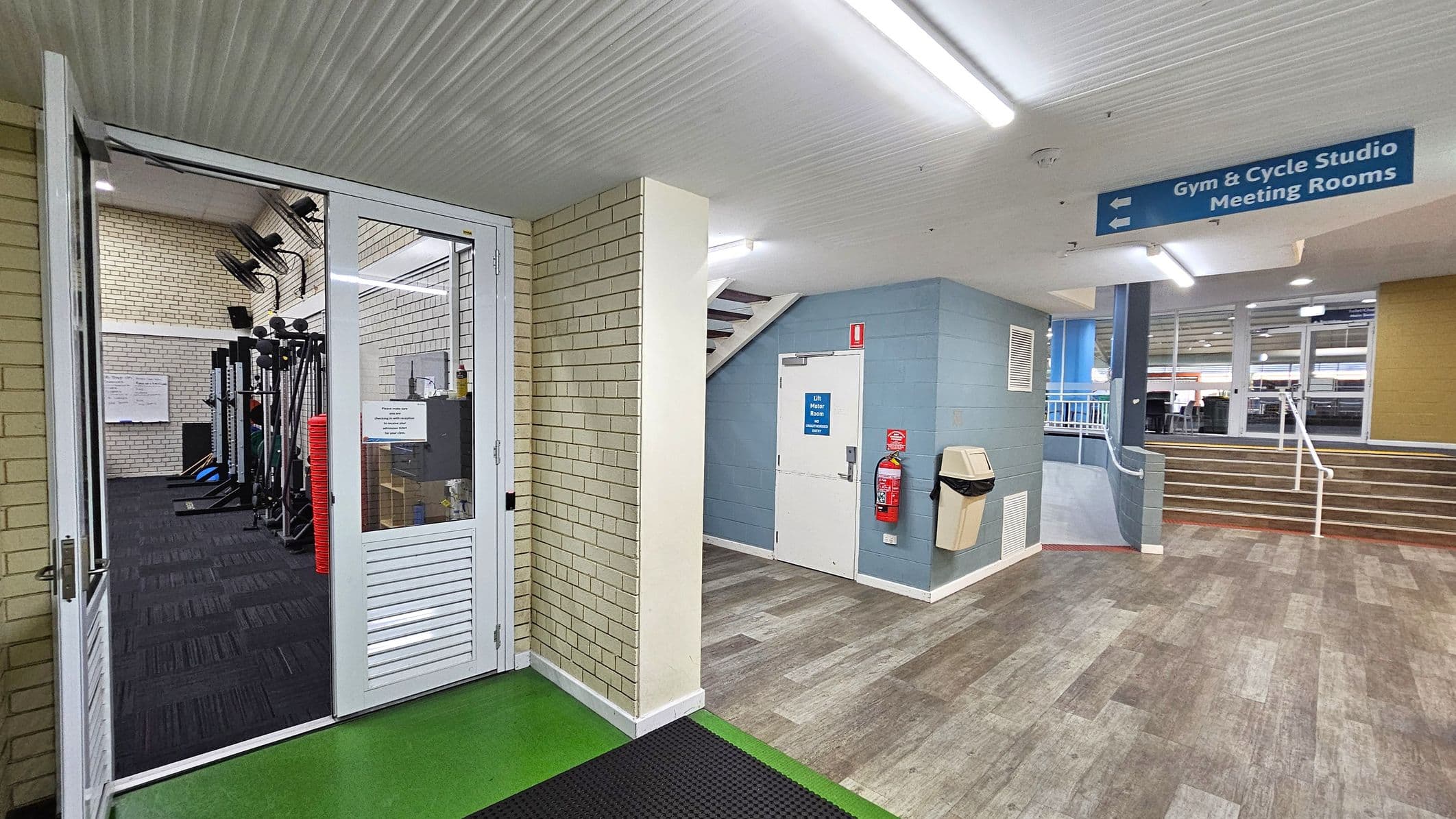 A gym entrance is on the left with glass doors and visible exercise equipment inside. The floor is green near the entrance and transitions to a wood-like pattern in the hallway. A white door with a blue sign is in the centre, next to a fire extinguisher and a bin. Stairs with a white railing lead up on the right. The ceiling is white with fluorescent lights. A blue sign above points to the gym, cycle studio, and meeting rooms.