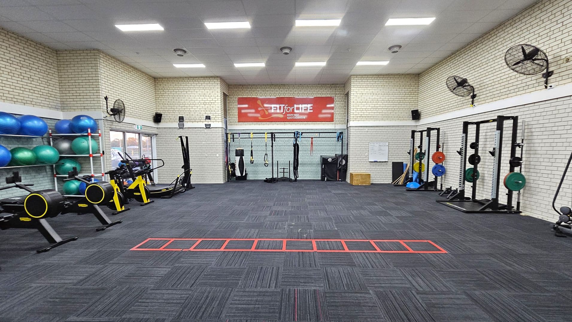 A gym with a dark carpeted floor and white brick walls. On the left, there are shelves with blue and green exercise balls and two rowing machines with yellow accents. In the centre, a red sign reads "FIT for LIFE" above various hanging exercise equipment. On the right, there are weightlifting racks with colourful weight plates. Ceiling fans are mounted on the walls, and the ceiling has bright rectangular lights.