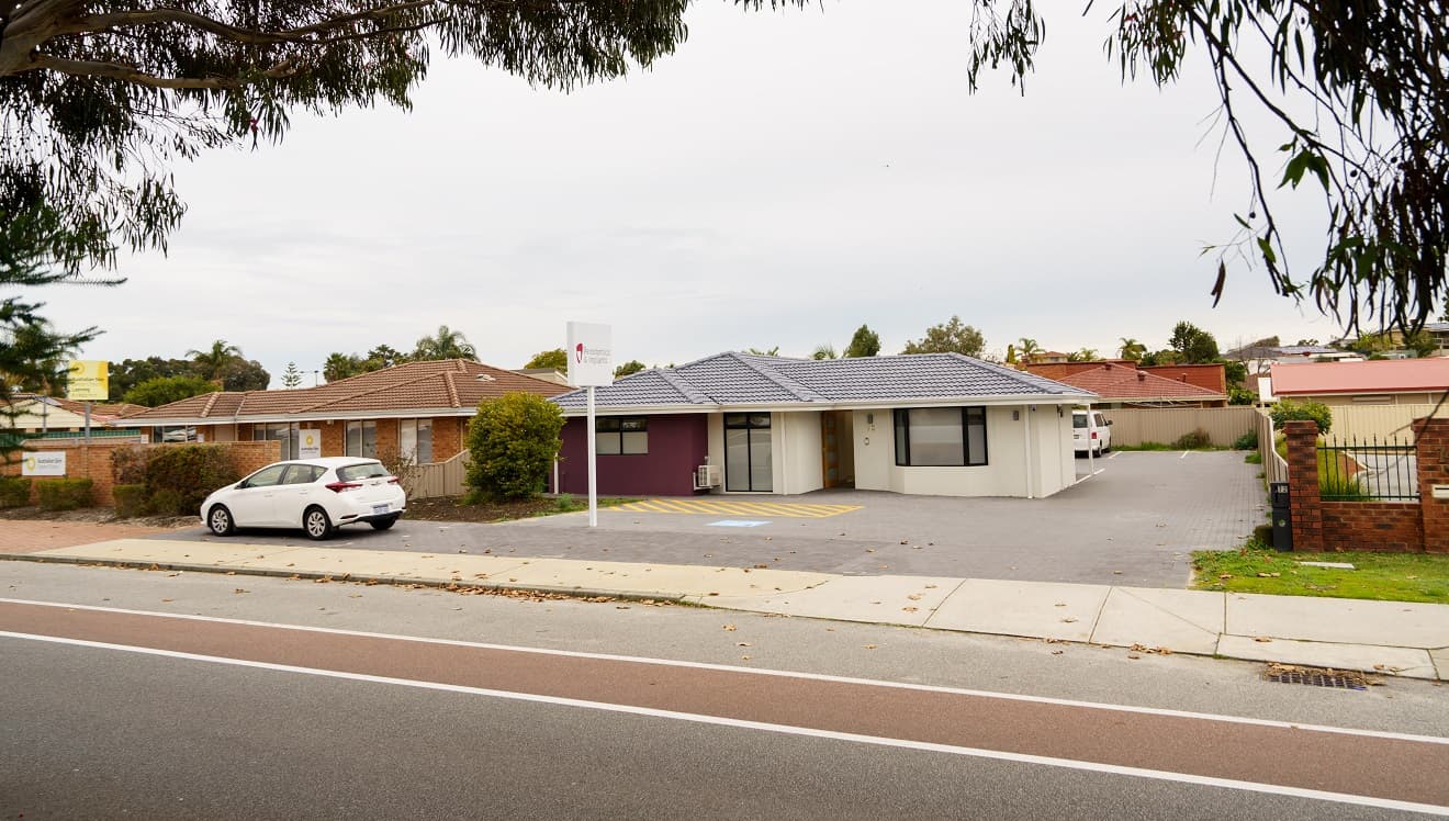 A cream building with a grey, tiled roof with a feature wall to the left in purple. The front door is timber with four glass panels. To the left is a white sign with 'Periodontics & Implants'. To the right is a glimpse of a small, white van parked behind the building and a sall white vehicle is parked out the front of the building. An accessible car parking space is directly adjacent to the front entrance. Other suburban style houses and businesses surround the business. Trees frame the view.