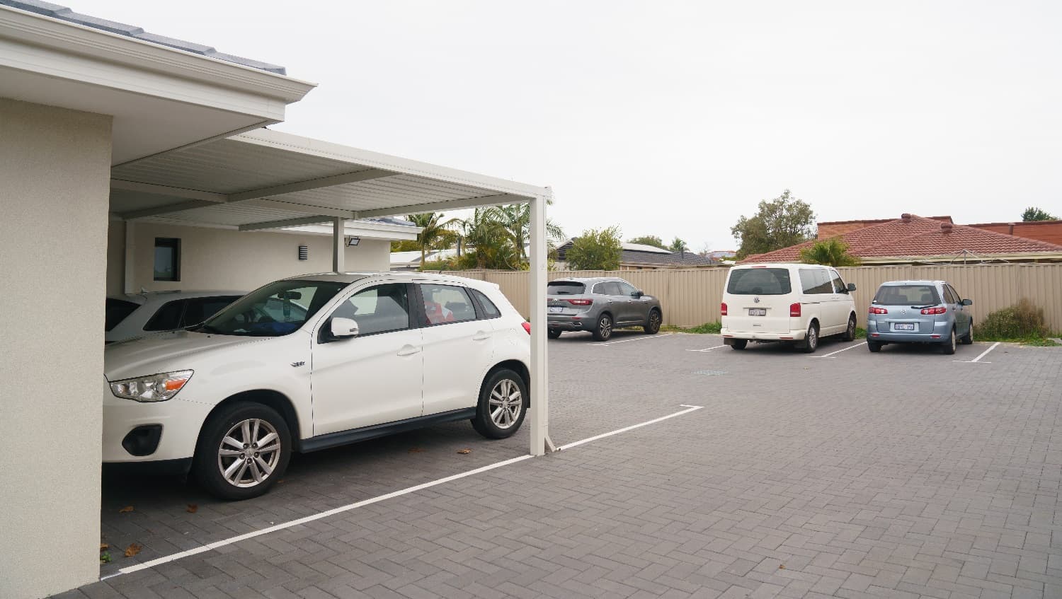 To the left are two white vehicles parked underneath a carport at the back of the cream building. Adjacent are is a small grey vehicle, a white van and a small silver hatchack parked up against the back fence. A glimpse of the house behind is visible and features a red tiled roof. Trees are including palm trees are visible in the backyard of the house located at the back of the business.