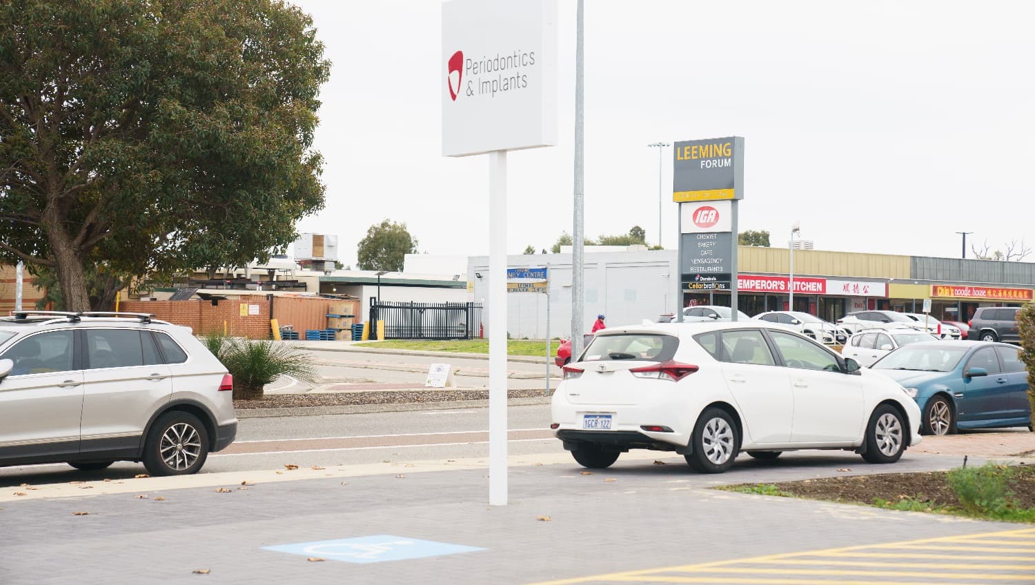 A silver vehicle is parked to the left on the roadside adjacent to a tree, whilst to the right a small white hatchback is parked outside the business underneath a white sign stating 'Periodontics & Implants'. Across the road a number of business are located at the 'Leeming Forum' including an IGA, Emperoros Kitchen and Noodle House. Vehicles are parked outside this retail precinct.