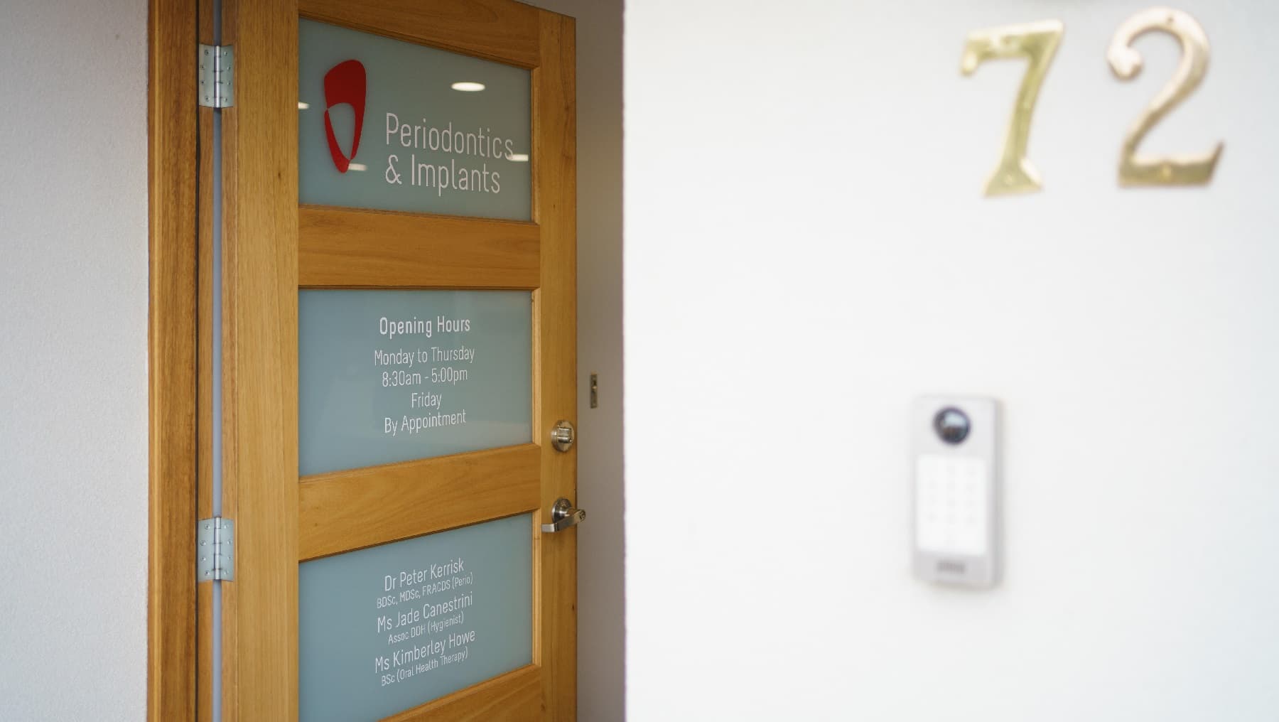 A close look of the front door open which is a timber door with three glass panels visible. The tope panel states 'Periodontics & Implants', the second panel down states the opening hours of Monday to Thursday of 8.30 am to 5pm and Friday by Appointment. The third panel down details the details of the dentist and assistants as well as their credentials. To the right is the number 12 with a door bell underneath this number.