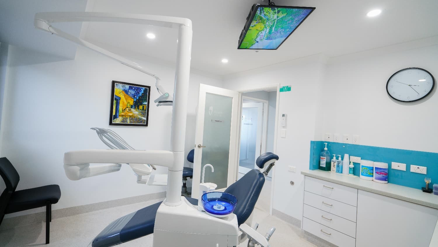 The clinic is neat and organized, with white walls and a sky-blue feature wall; the clinic has four drawers near the door. There is a reclining examination chair that's dark blue in color. This chair appears comfortable and is attached to the basic equipment used by dentists/periodontists such as chairside spittoon, suction, the LED lamp and the like. There is a LED TV mounted in the ceiling above and a painting posted on the wall that used bright and vibrant shades of blue and orange.