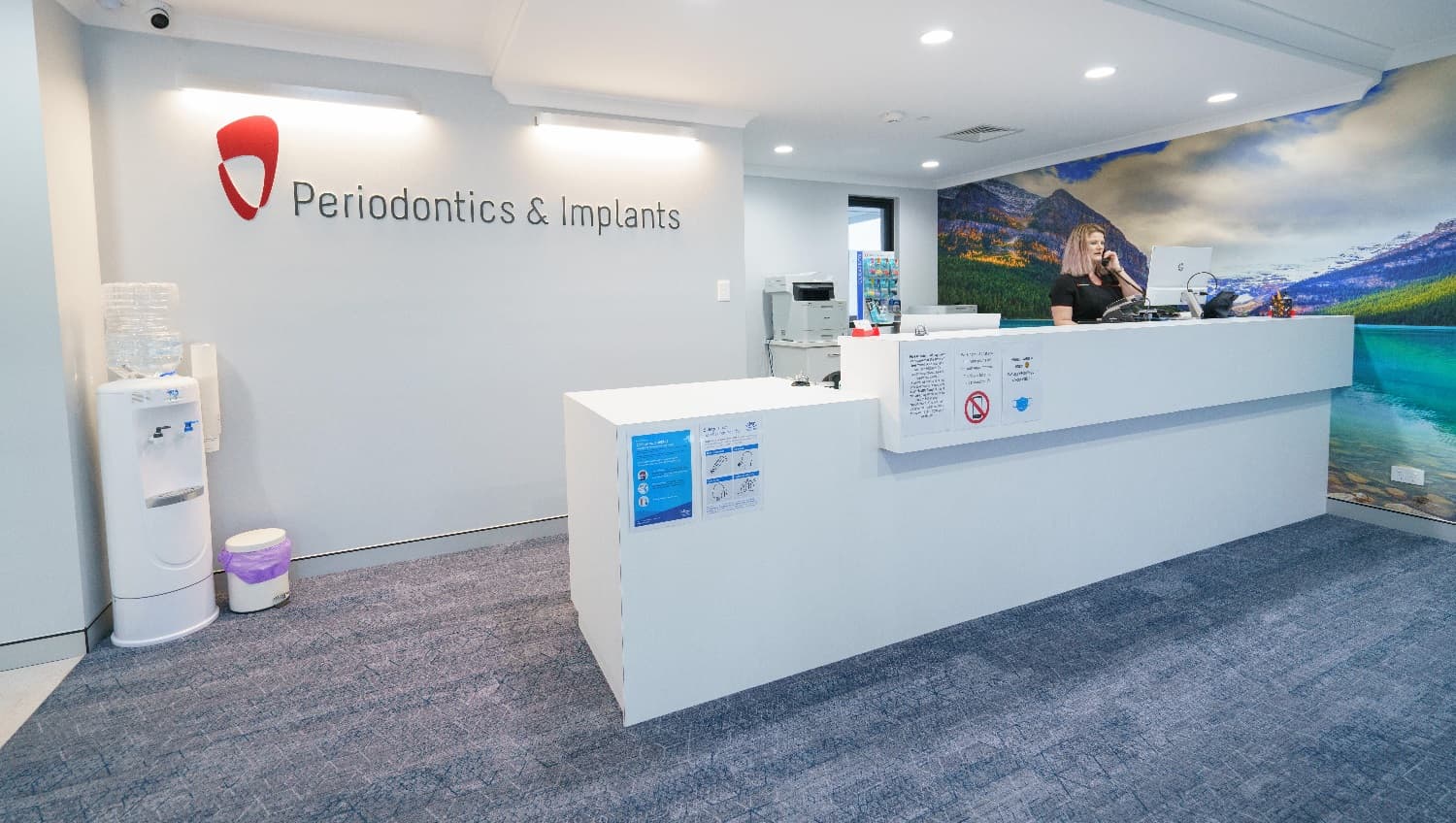 The long reception desk is painted white, the entire floor is carpeted, a large wall further to the right has a landscape picture of clouds, mountains, and water that adds a cozy feel to the room, and a white cabinet is placed at the back corner of the long desk while securing a space between them, to give more space for the clinic personnel. The clinic’s red logo is highlighted as its background wall is white. A desktop computer and telephone unit for use by the staff are on the desk.