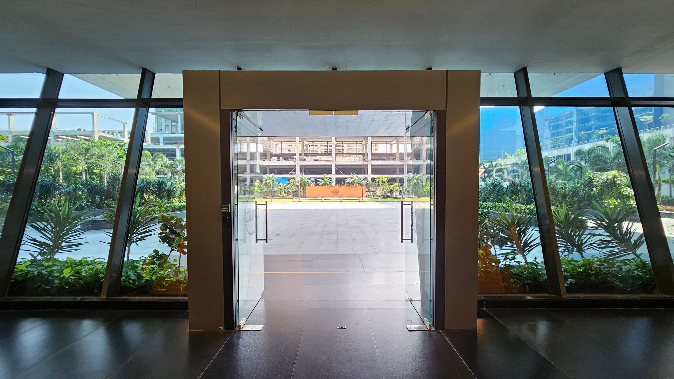 A glass door in the centre opens to an outdoor area with a tiled floor. On either side, large glass panels provide a view of lush green plants and trees. The background features a modern building with multiple floors and a clear, structured design. The floor inside is dark and glossy, while the ceiling is plain and light-coloured.