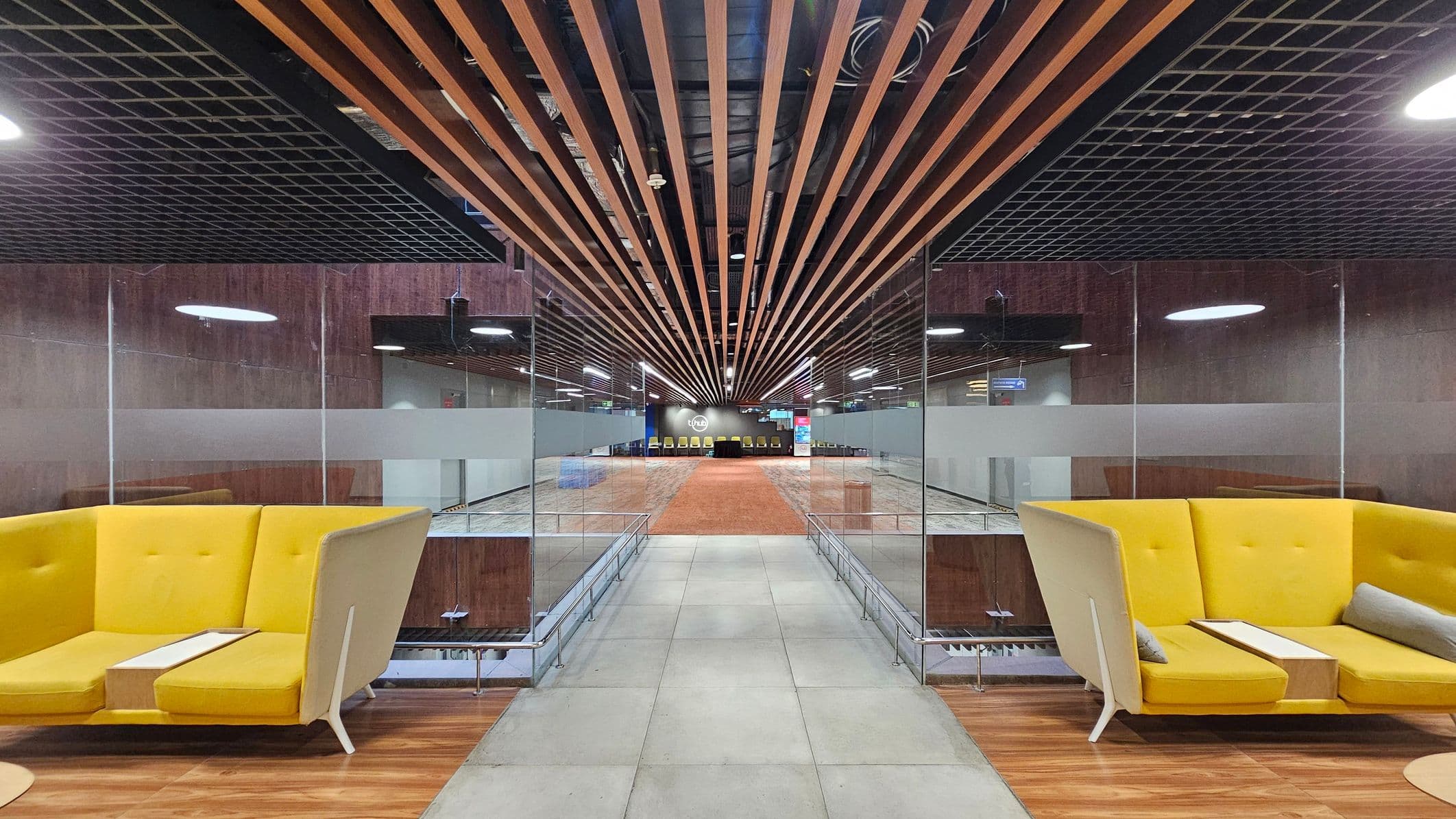 A modern interior with a symmetrical design. On the left and right, there are yellow, high-backed sofas with small tables attached. The floor is a mix of grey tiles and wood. The ceiling features a grid pattern with wooden slats running lengthwise. In the centre, a glass partition separates the seating area from a spacious hallway leading to a distant wall with a logo and a row of chairs. The lighting is bright, with circular fixtures on the ceiling.