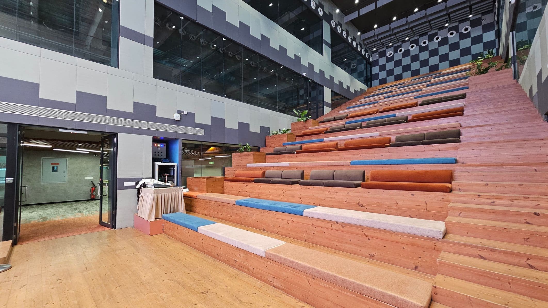 A modern interior space with a large, stepped seating area on the right, featuring wooden benches with colourful cushions in shades of blue, orange, and brown. On the left, there is a glass entrance with a door leading to another room. Above the entrance, the wall is decorated with a geometric pattern in grey tones. The ceiling is high with visible lighting fixtures and a checkered pattern on the upper wall. The floor is wooden, matching the seating area.