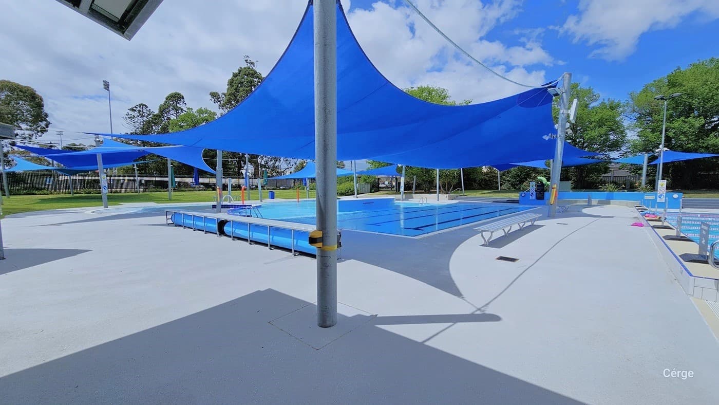 Heated leisure pool and playful water features with blue sunshades. Each sunshade is tied around grey posts. On the sides of the heated leisure pools and water playful features are metal benches. The metal bench on the end of the heated leisure pool has a large blue circular roll of waterproof fabric. Wrapped around the grey post in the middle part of the right pool deck is a yellow floating device.