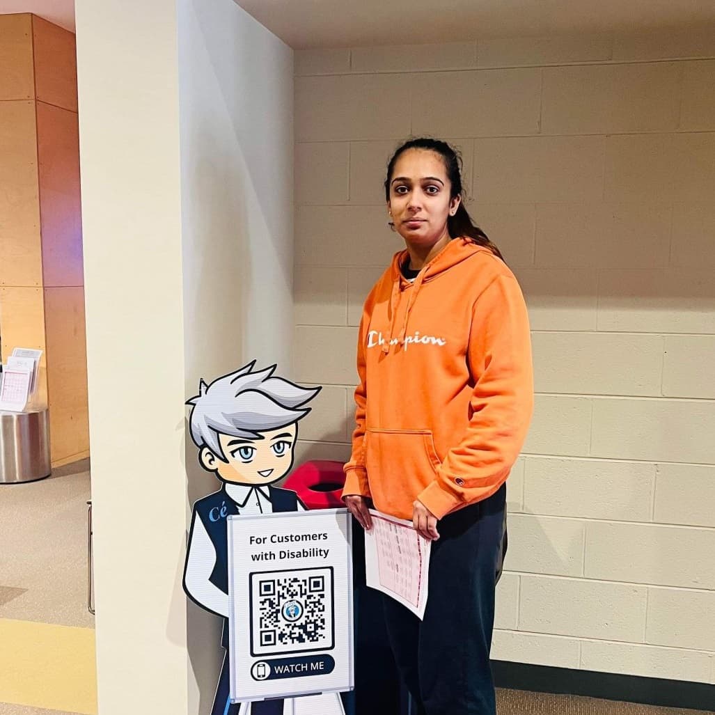 A woman in an orange hoodie stands next to a Cérge cartoon cutout with a QR sign. A woman in an orange hoodie stands next to a Cérge cartoon cutout with a QR sign.